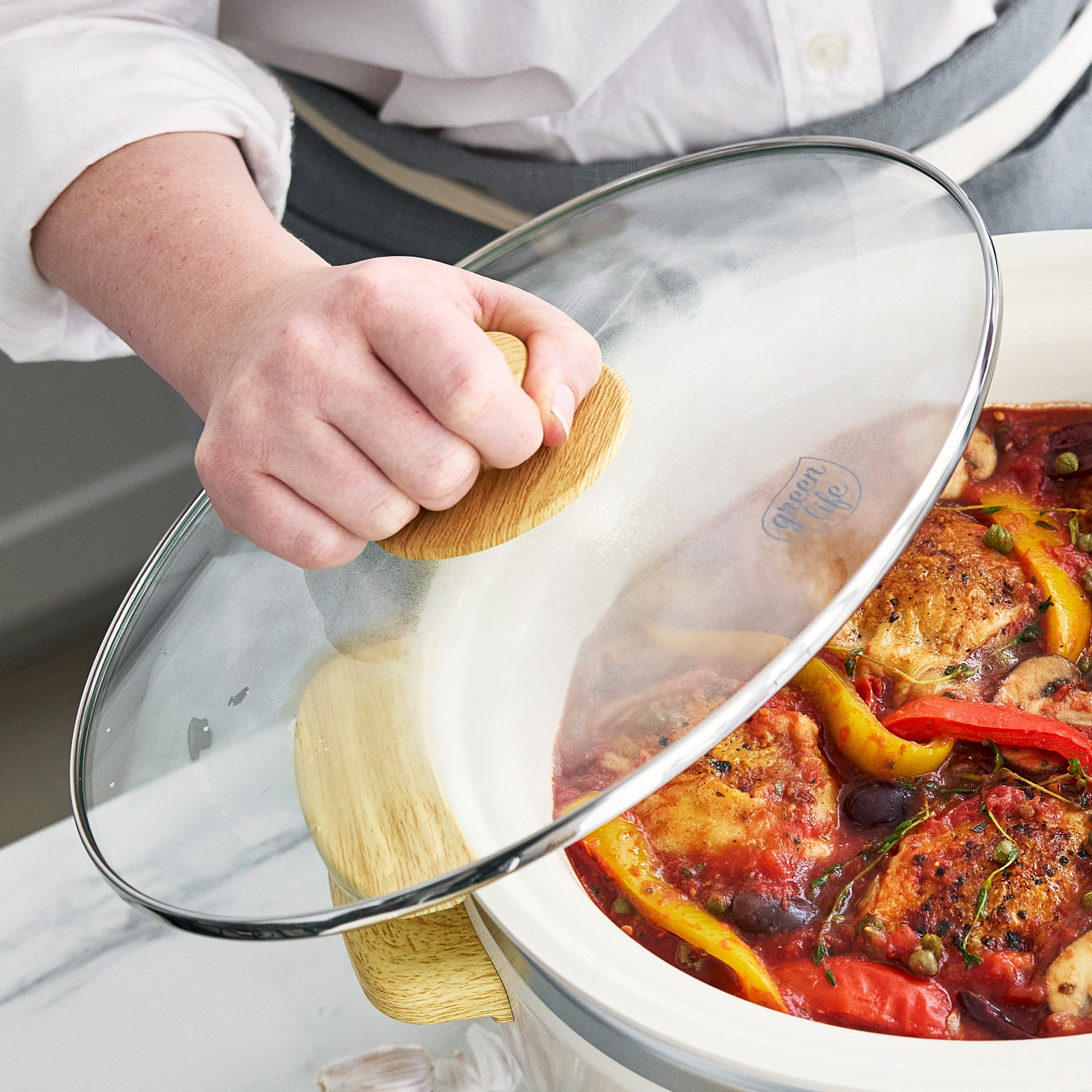 Person holding a glass lid on a dish of cooked chicken and vegetables.