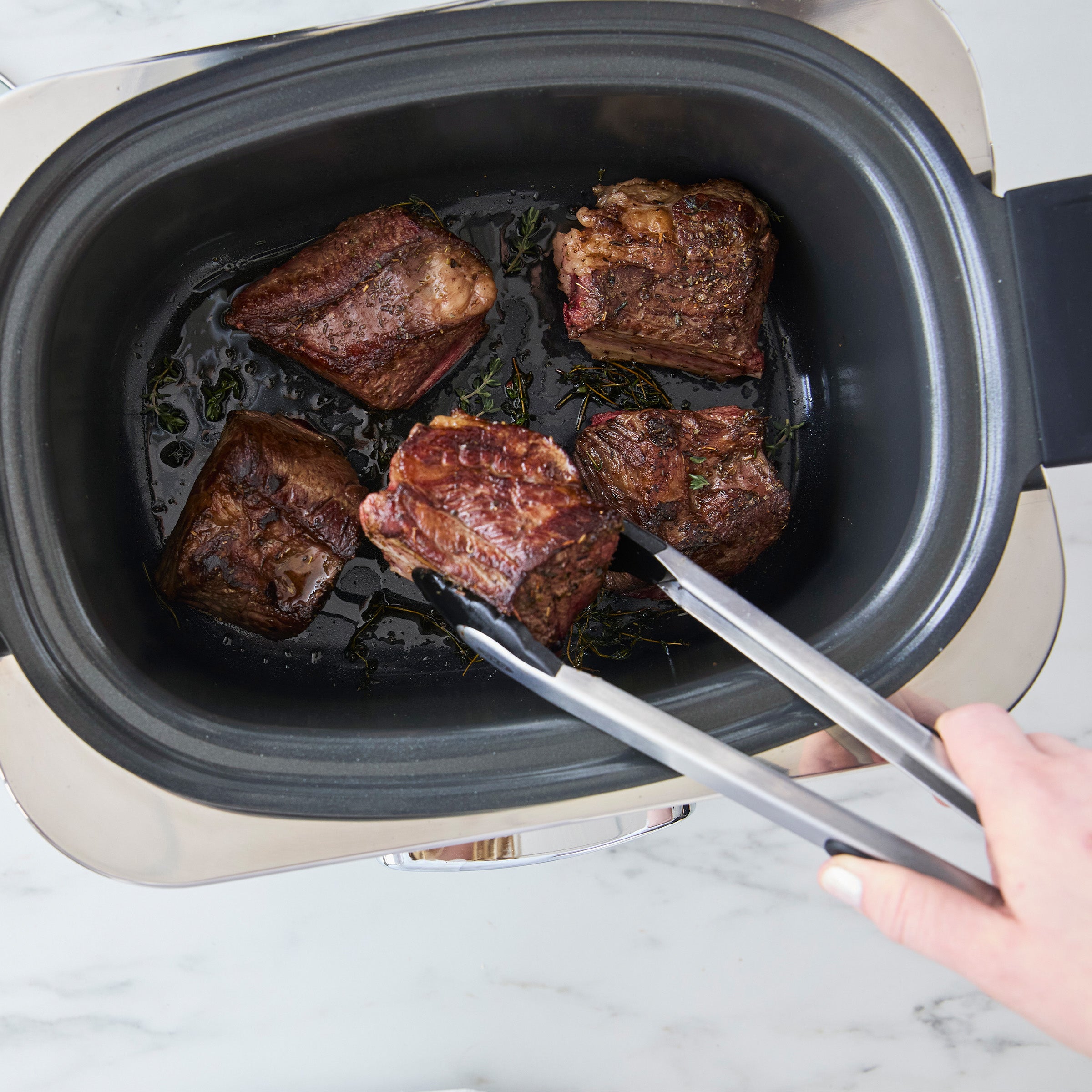 Cooking meat in a slow cooker with tongs on a marble surface