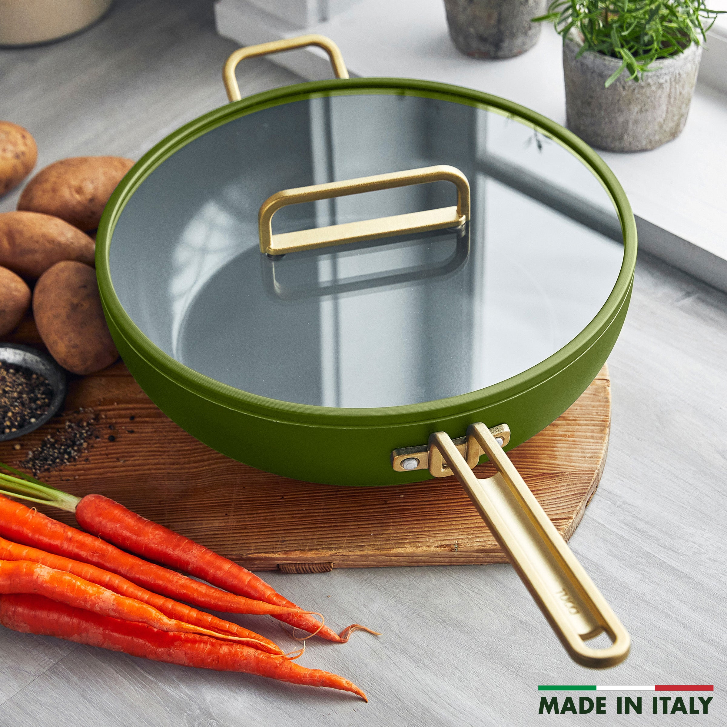 Green saute pan with gold handles on a wooden cutting board, surrounded by carrots and potatoes.