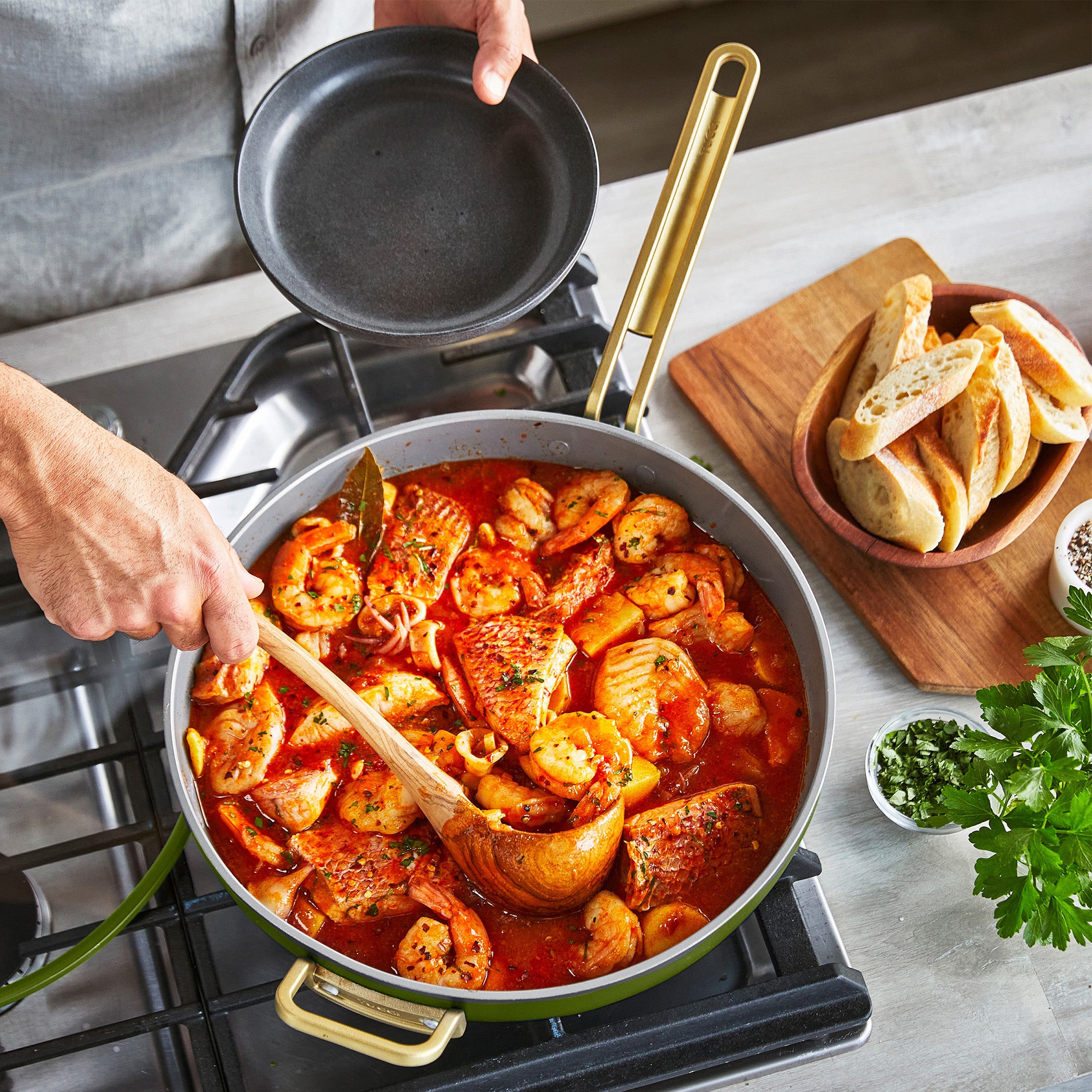 Person stirring a pan of seafood on a stove with a bowl of bread and herbs nearby.