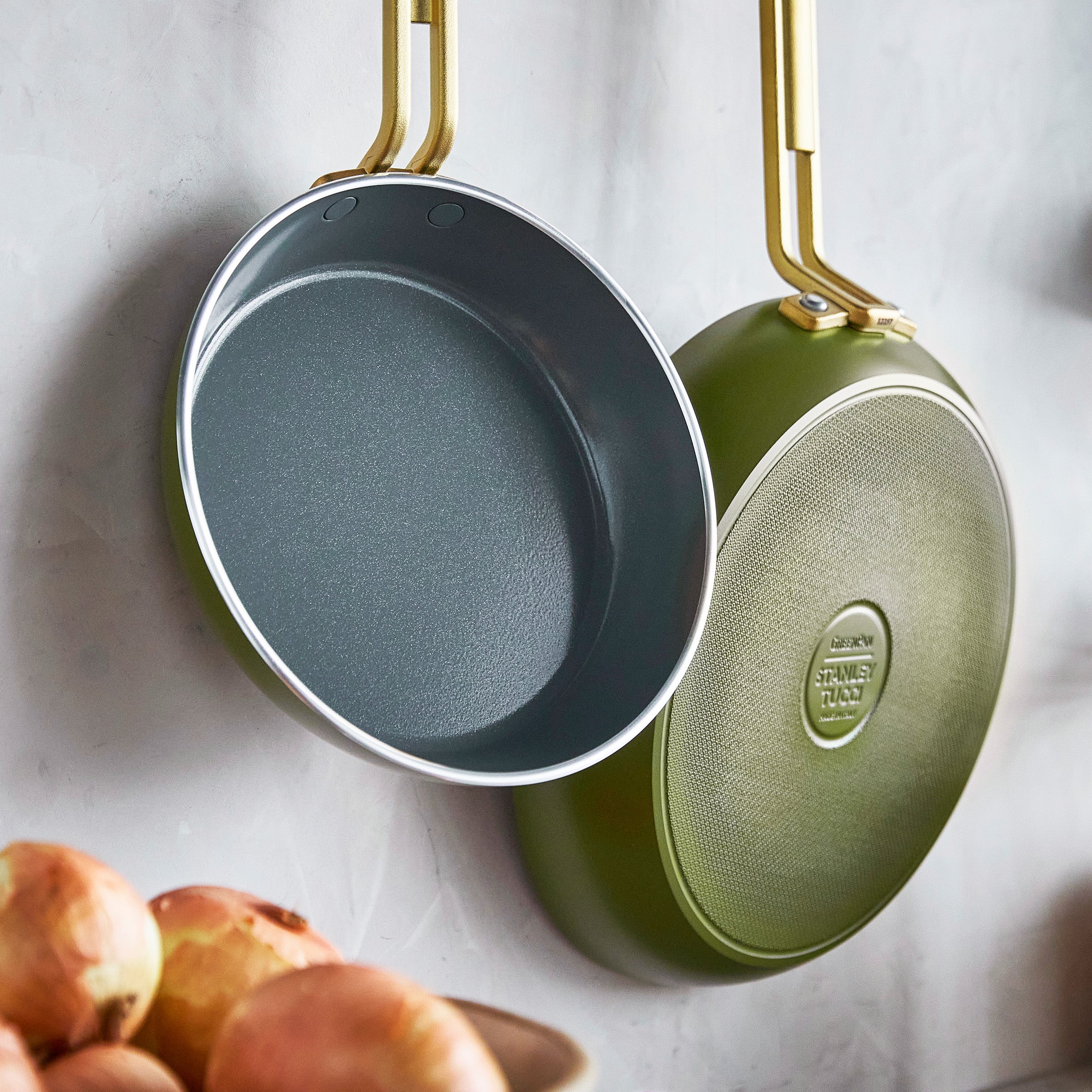 Two green frying pans hanging on a wall with onions below.