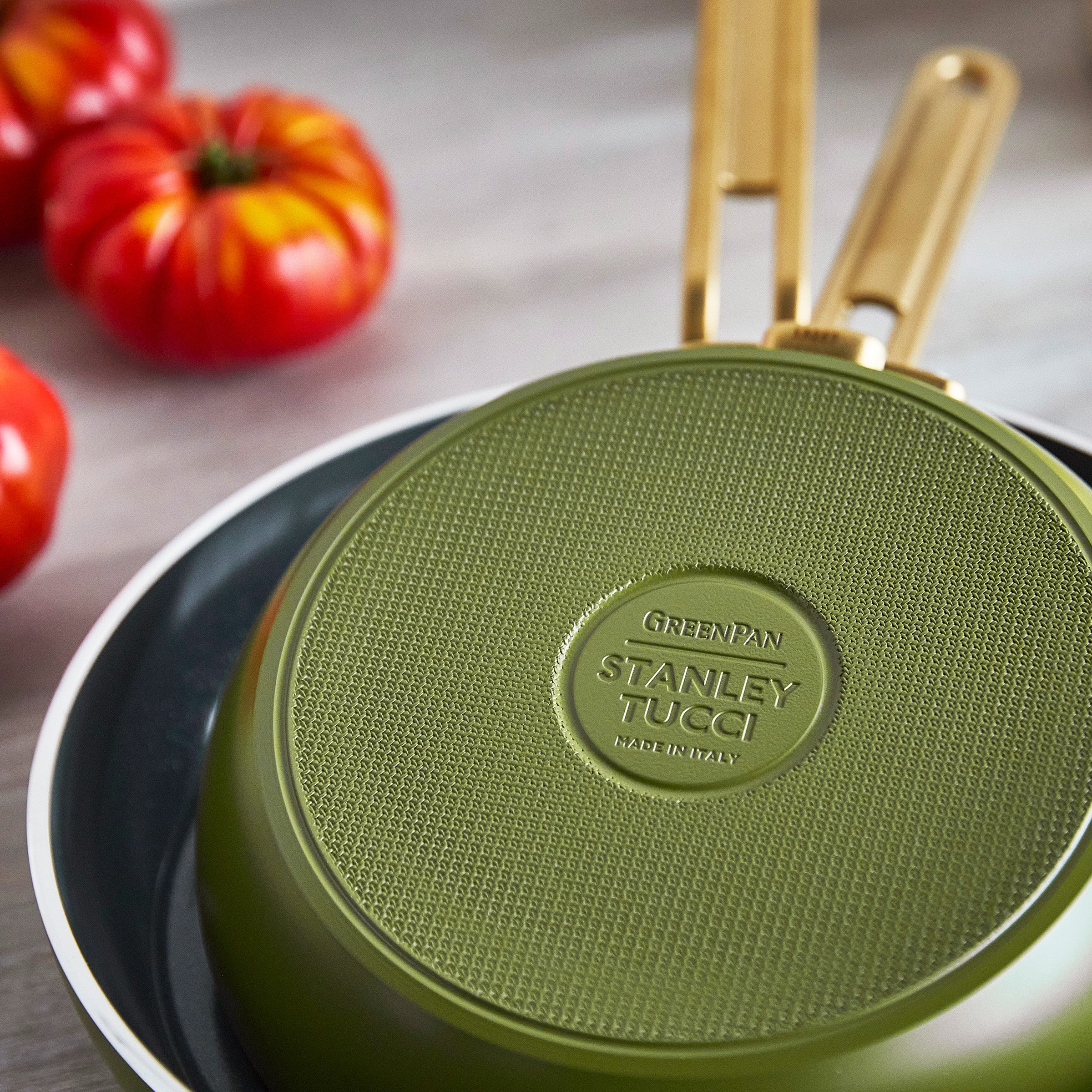 Green frying pan with Stanley Tucci logo on a wooden surface with tomatoes in the background