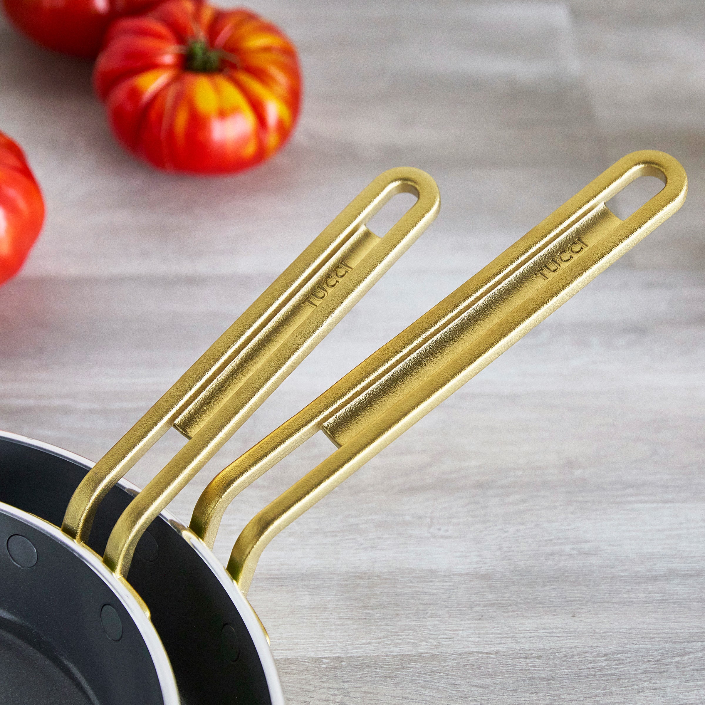 Two gold-handled frying pans on a light wooden surface with red tomatoes in the background.