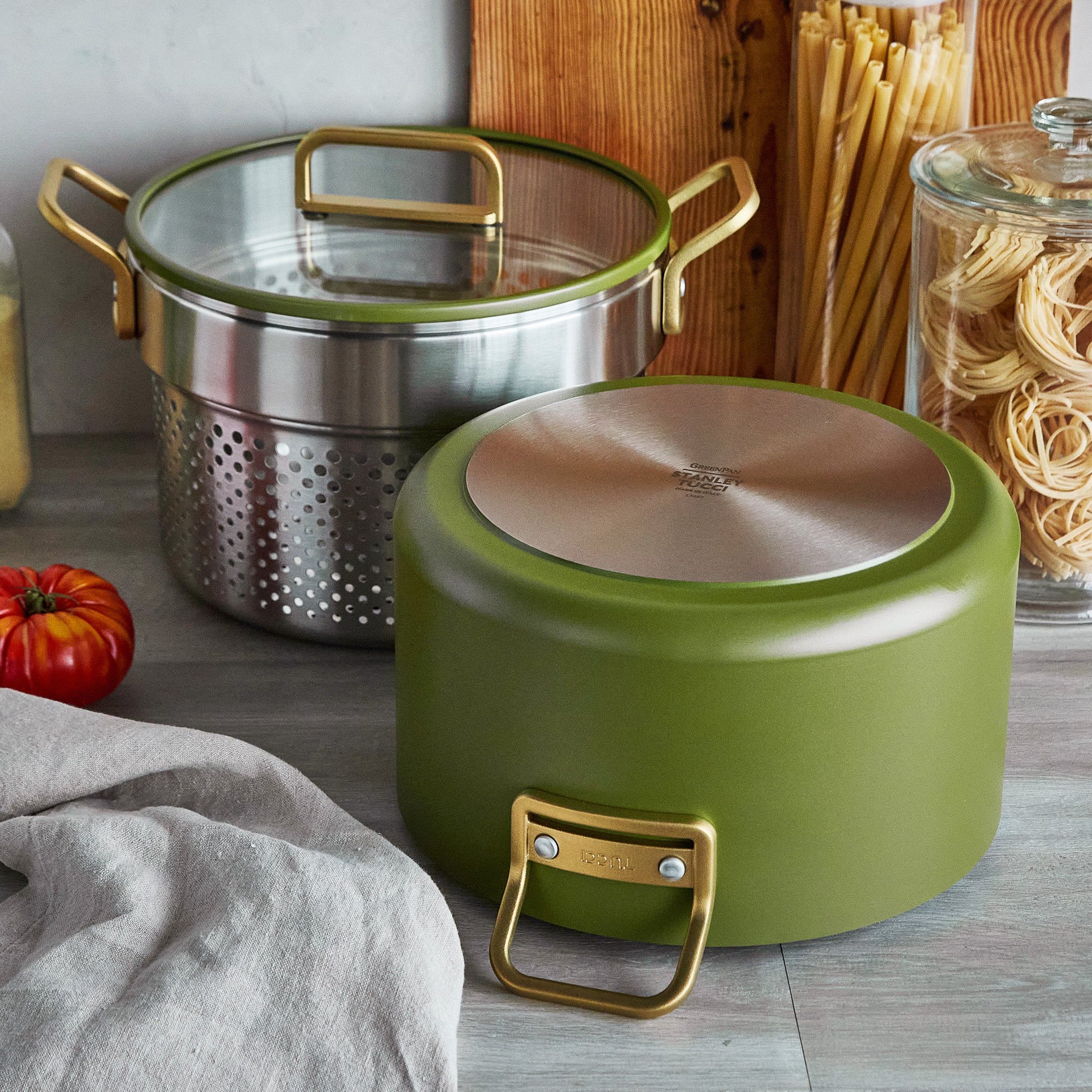 Green cookware on a kitchen counter with pasta and a tomatos in the background