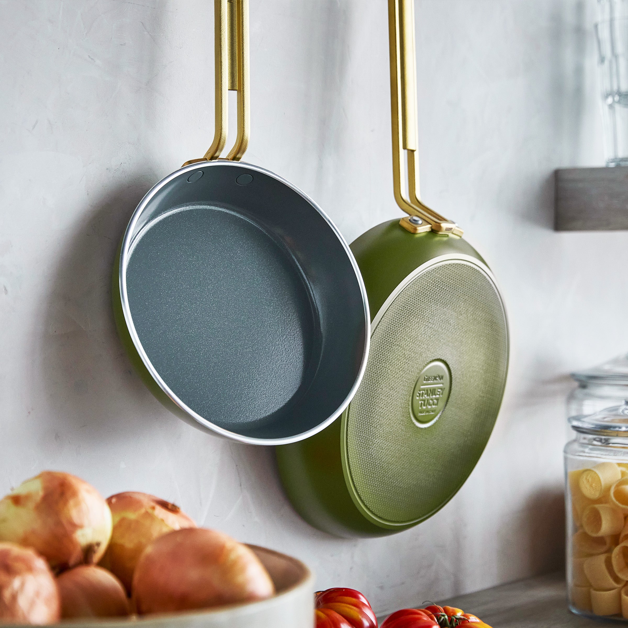 Two green frying pans hanging on a wall with ingredients below.