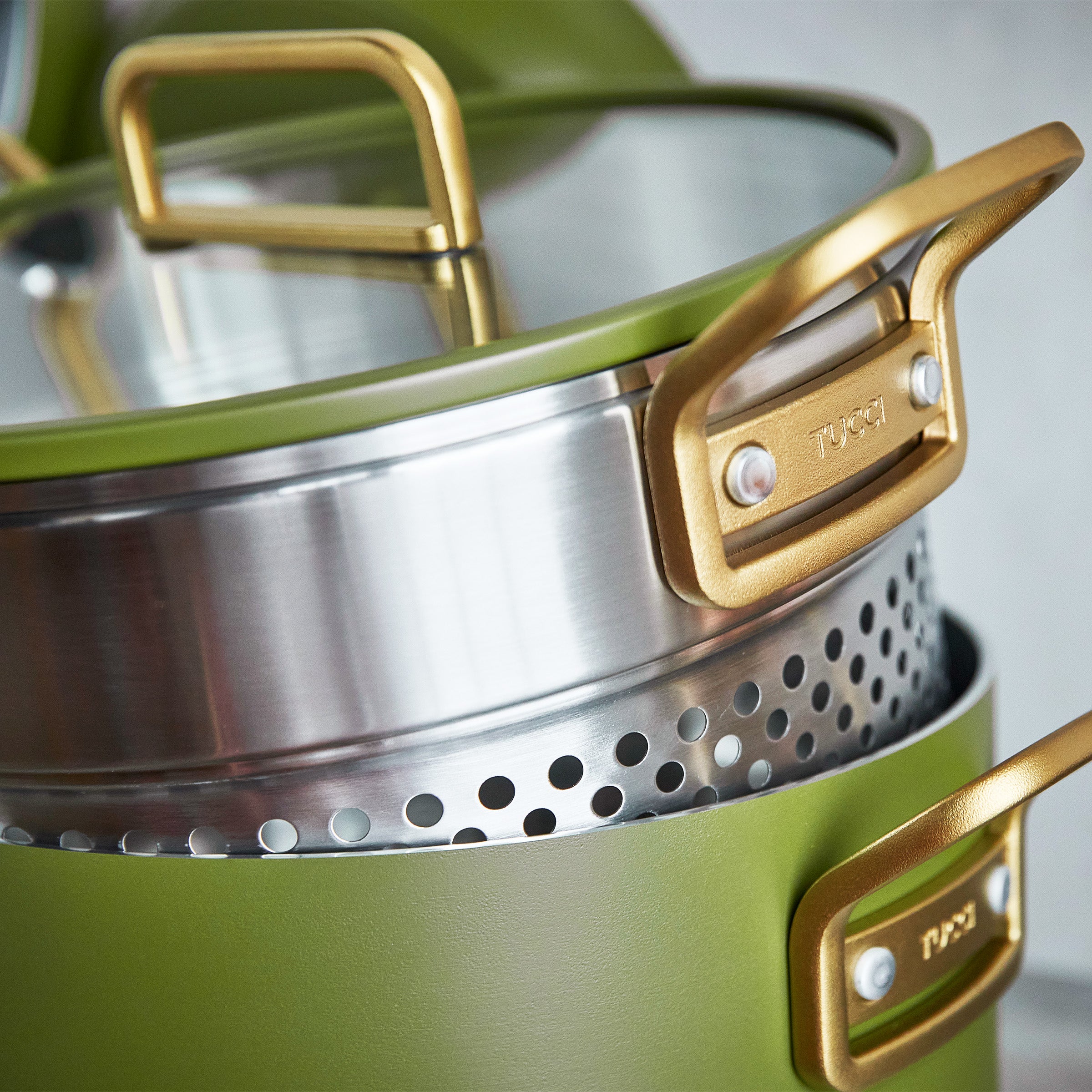 Green cookware set with gold handles and stainless steel steamer on a neutral background