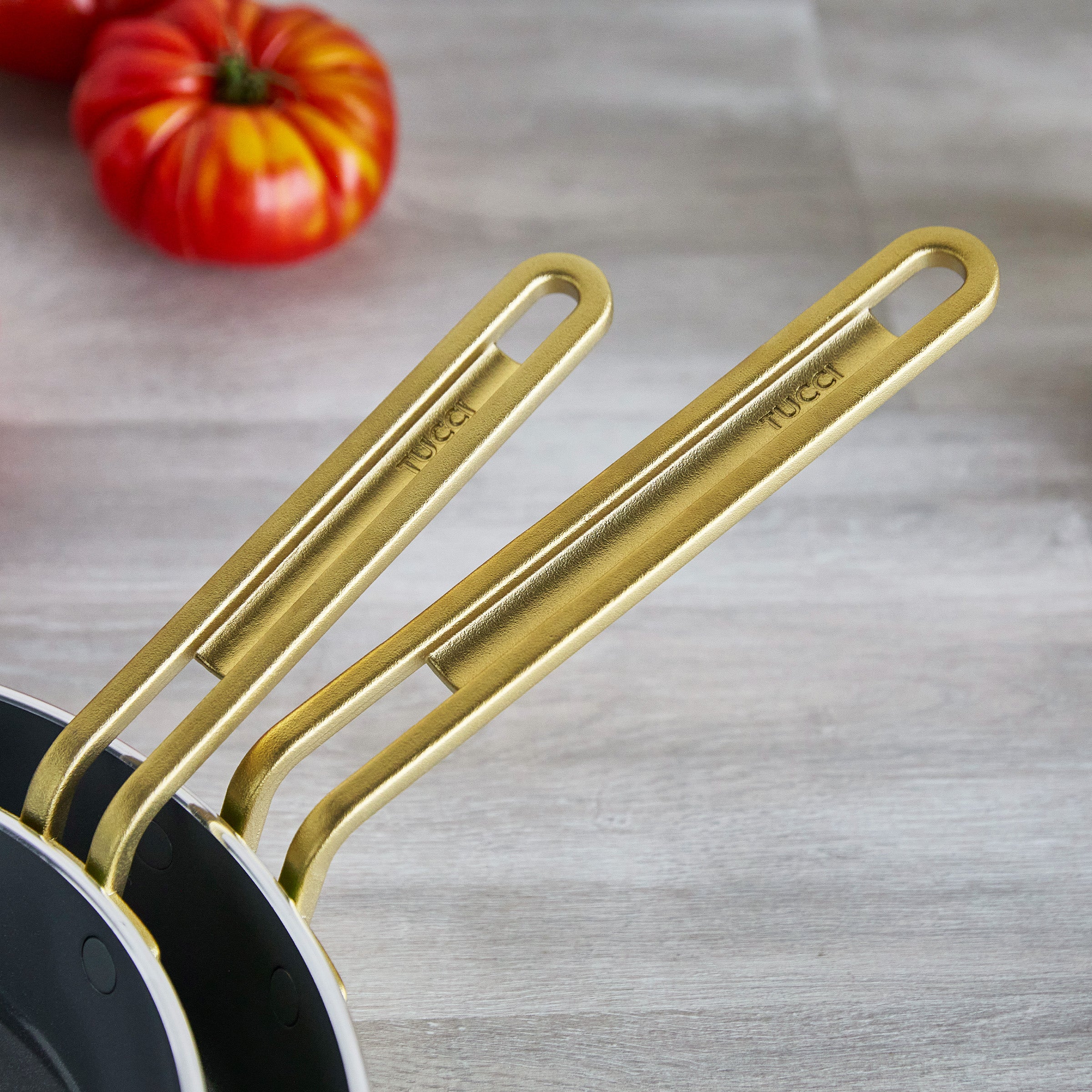 Two gold-handled frying pans with tomatoes in the background on a wooden surface