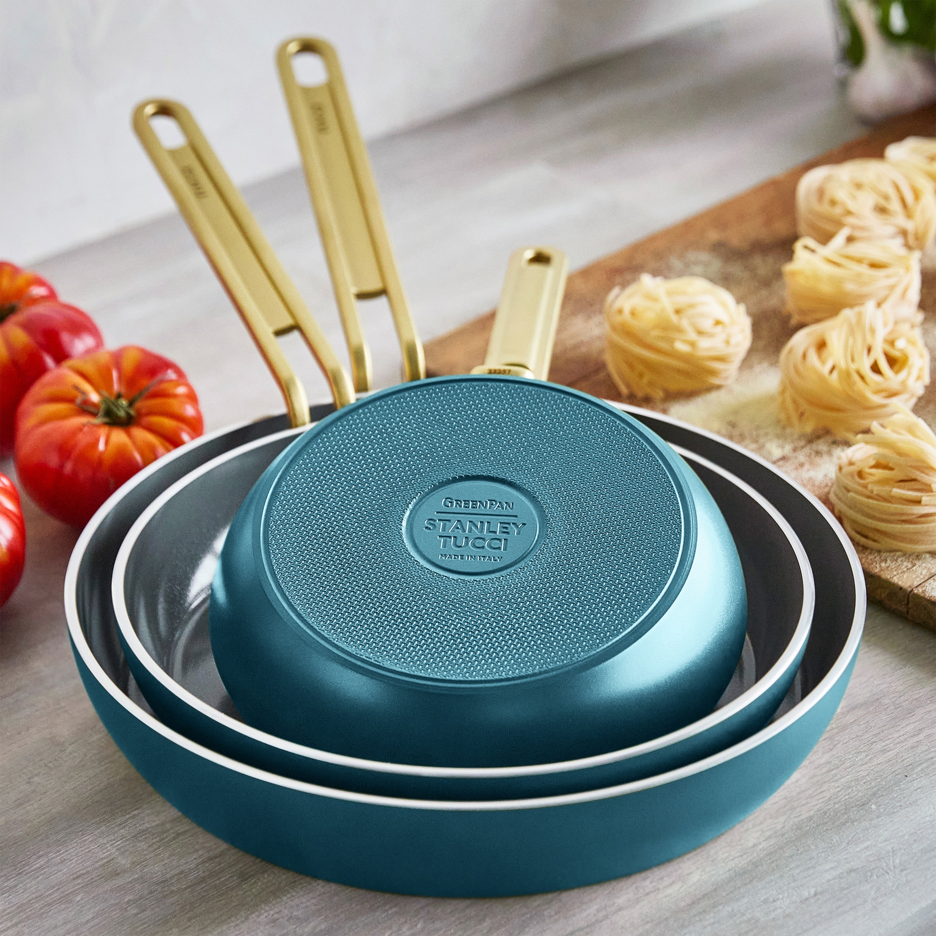 Set of teal frying pans with gold handles on a kitchen counter with tomatoes and pasta.