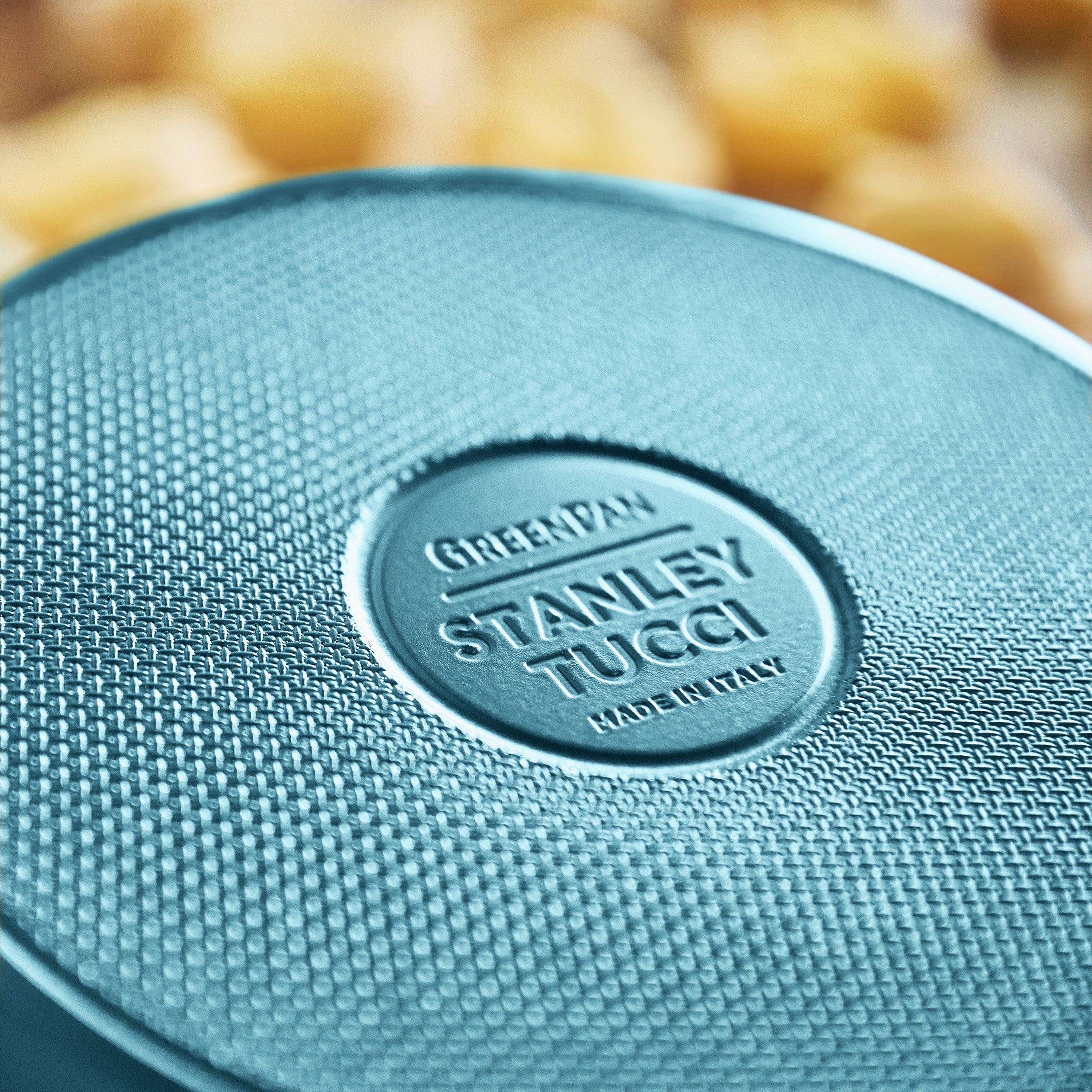 Close-up of a teal frypan with 'Stanley Tucci' branding on a blurred background.