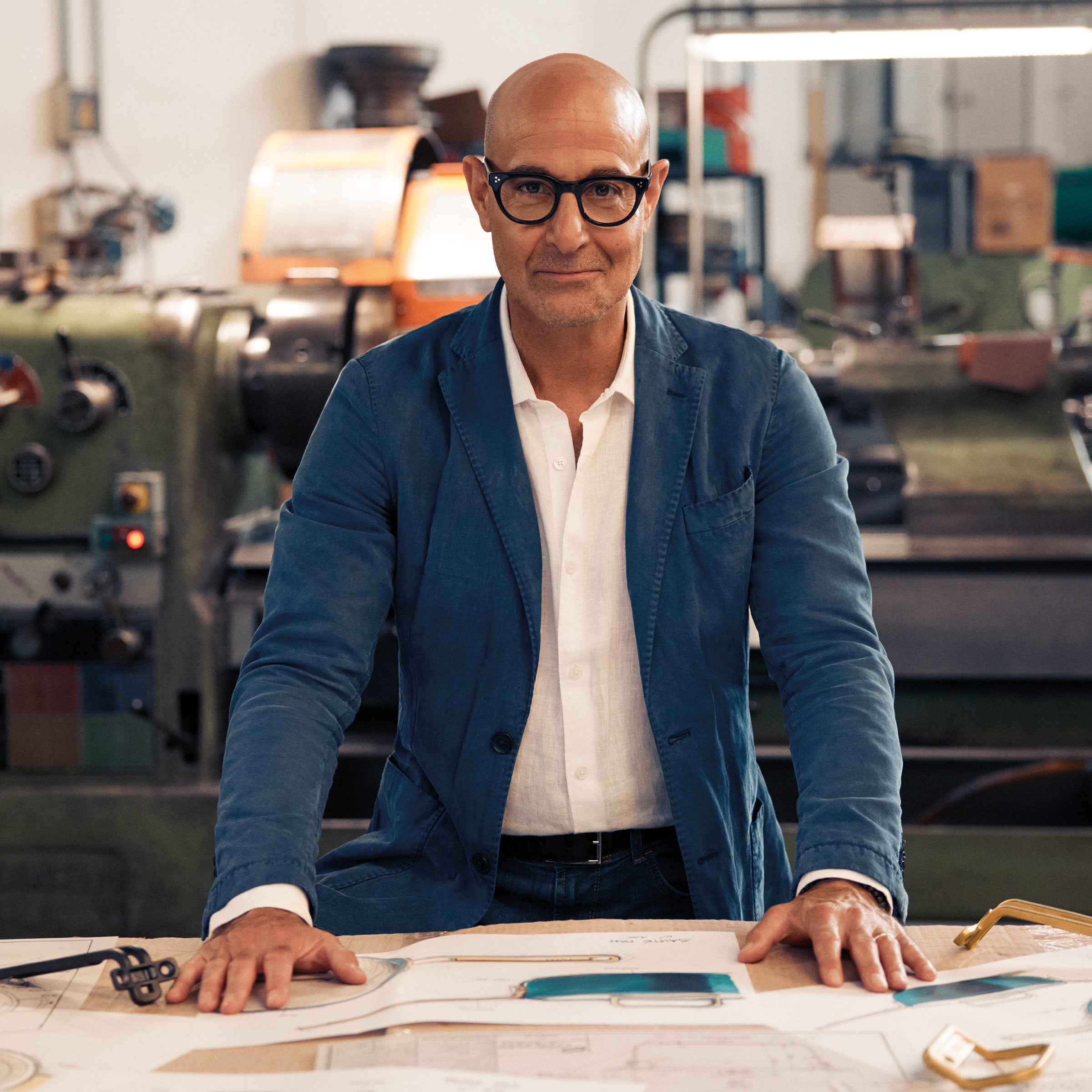 Stanley Tucci in a blue jacket and glasses standing in an industrial setting with machinery in the background.