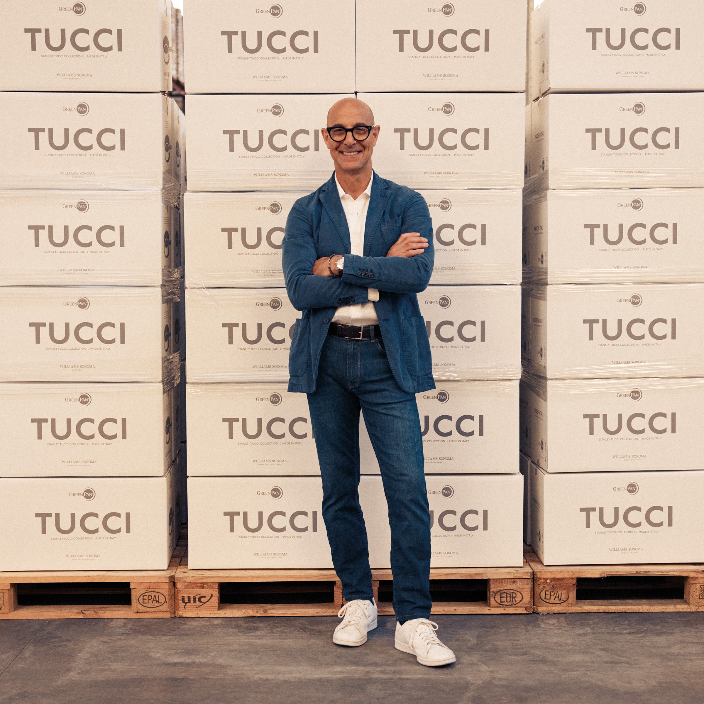 Stanley Tucci standing in front of Tucci cookware boxes, wearing a blue suit.