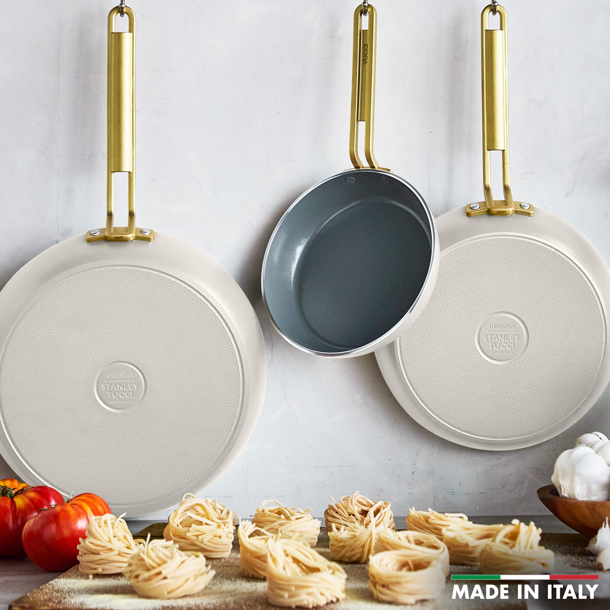 Three white frying pans with gold handles on a white background, with pasta and vegetables at the bottom.