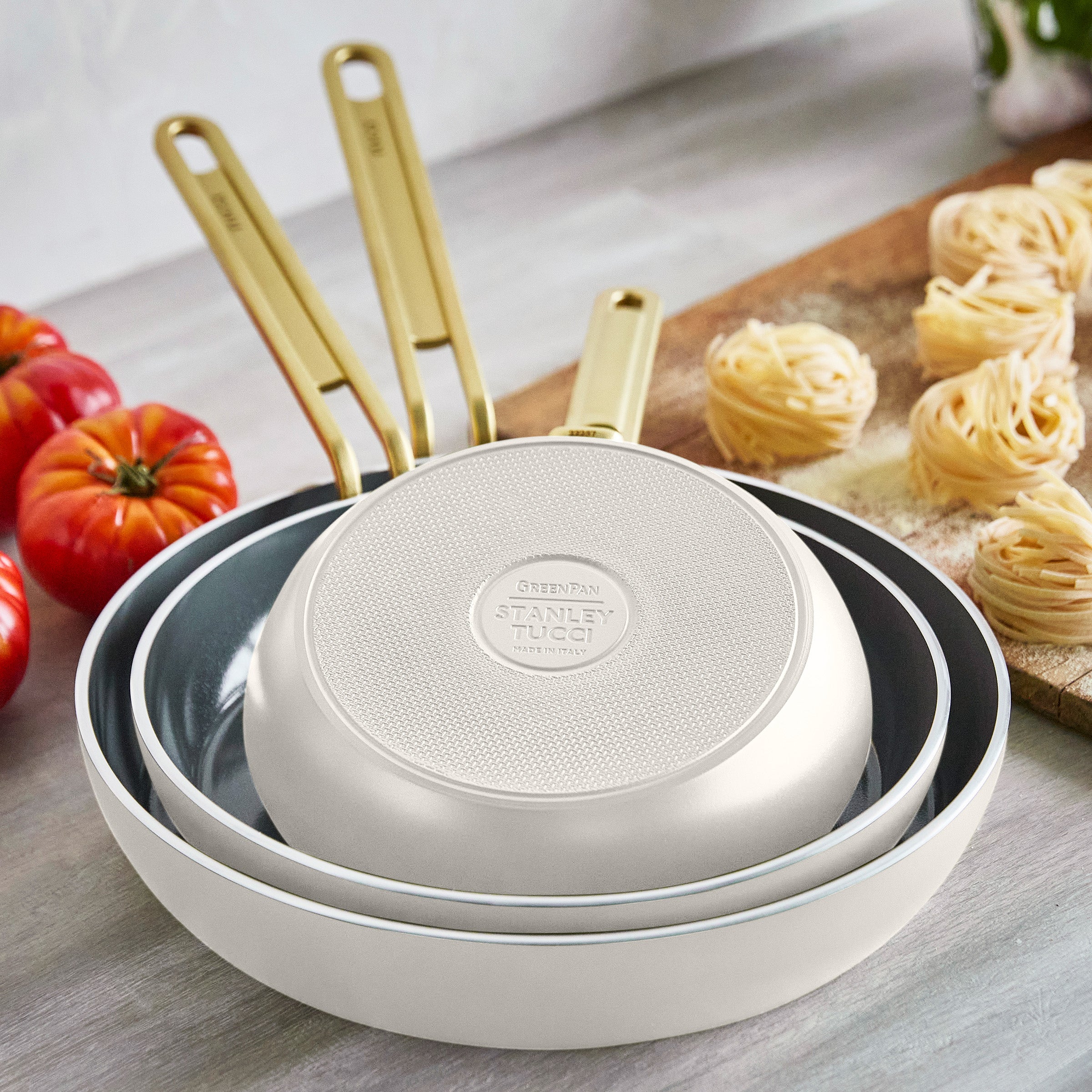 Set of white frypans with gold handles on a wooden surface with tomatoes and pasta.