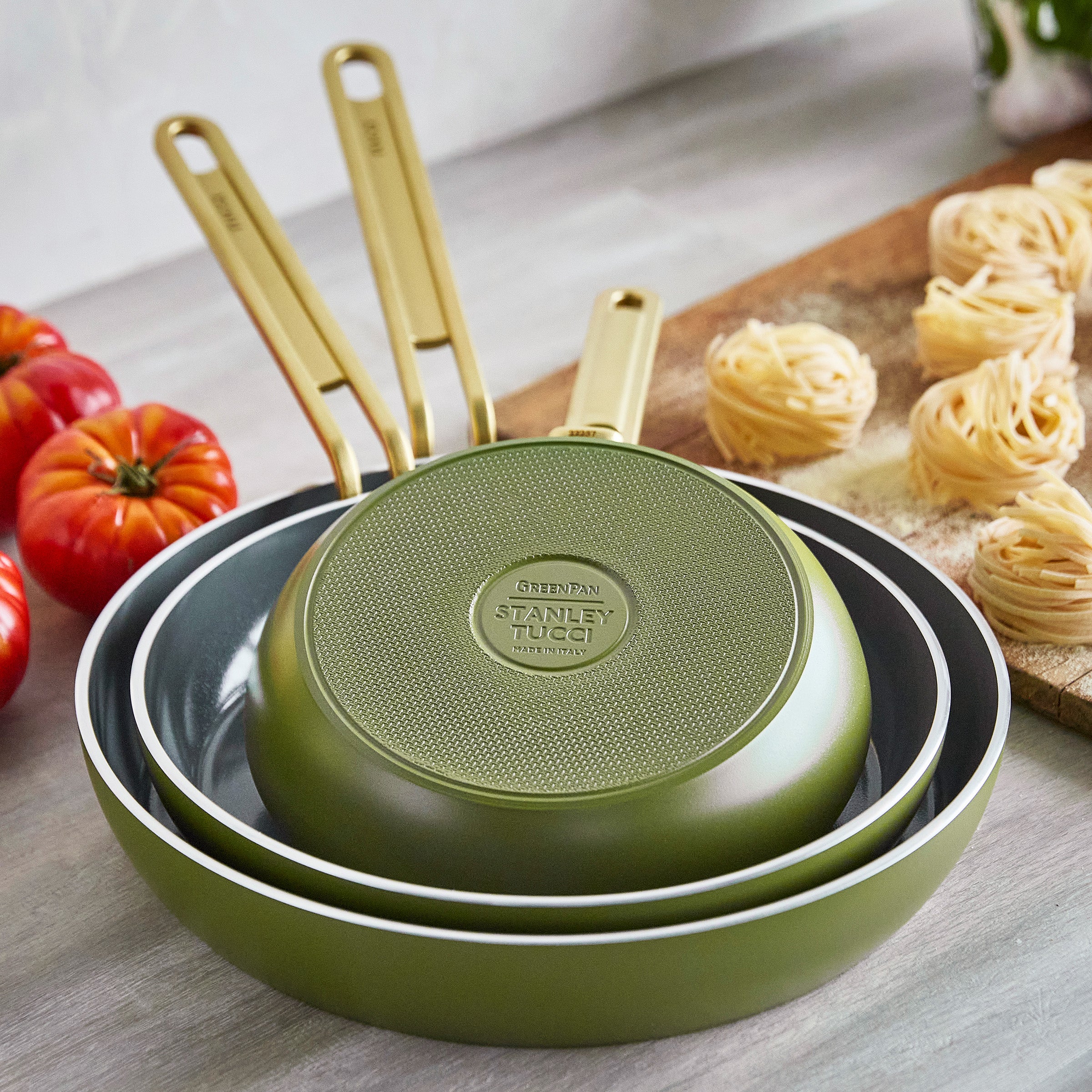 Set of green frying pans with gold handles on a wooden surface with tomatoes and pasta.