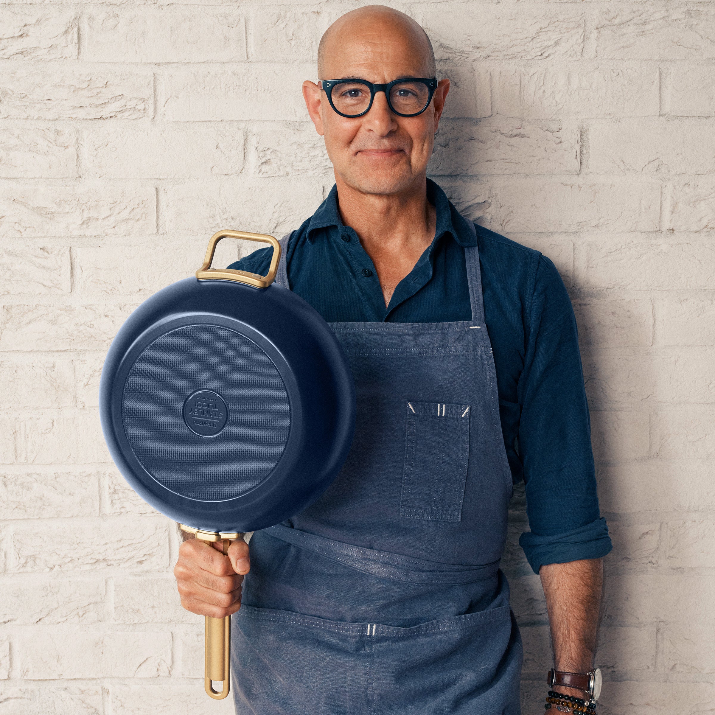 Stanley Tucci holding a blue saute pan against a light brick wall