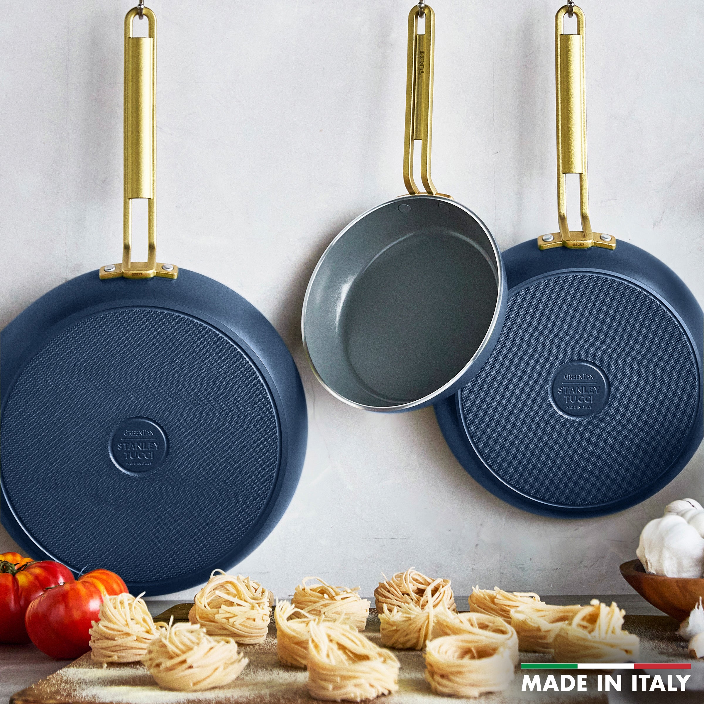 Three blue frying pans with gold handles on a white background, with pasta and tomatoes in the foreground.