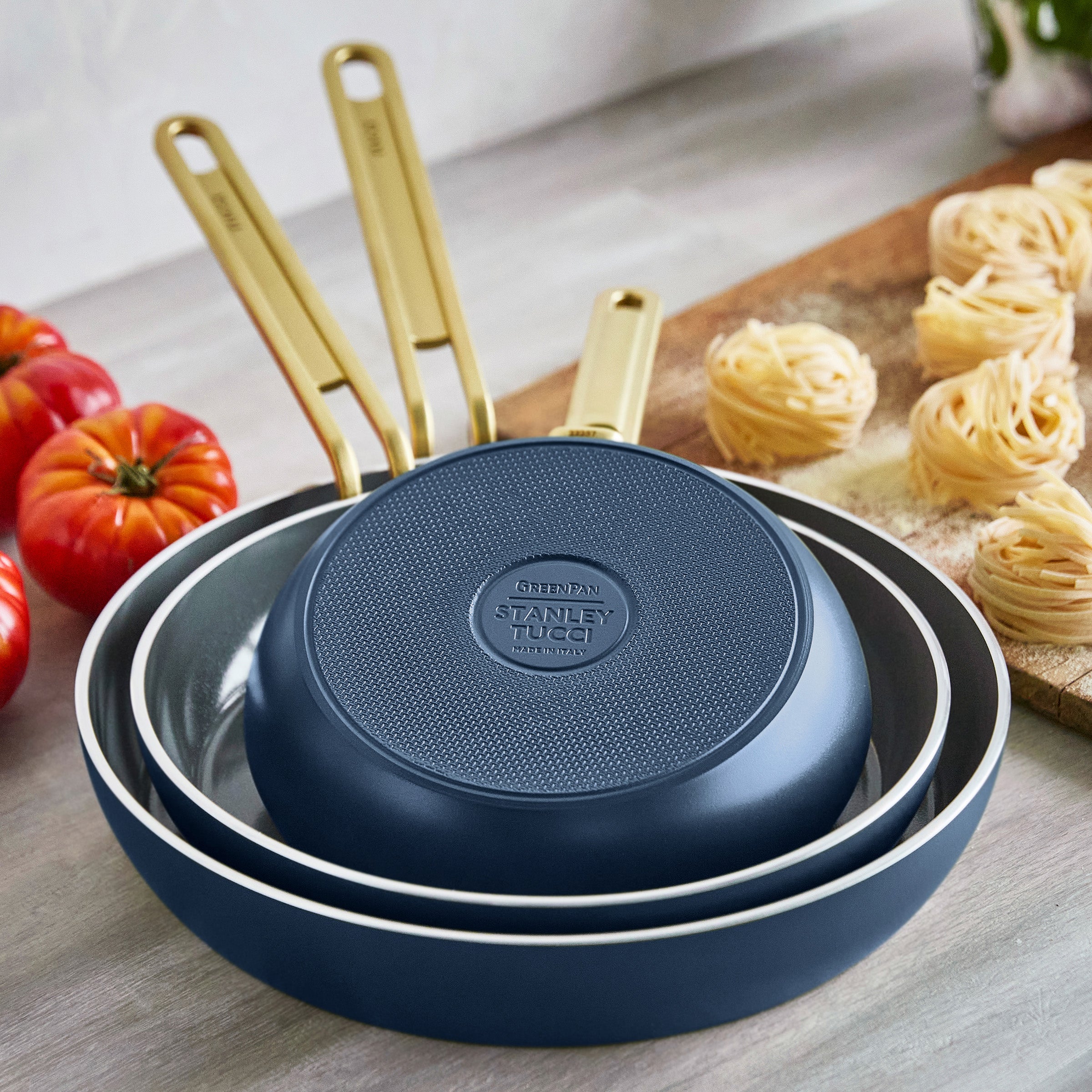 Set of blue frying pans with gold handles on a wooden surface with tomatoes and pasta.