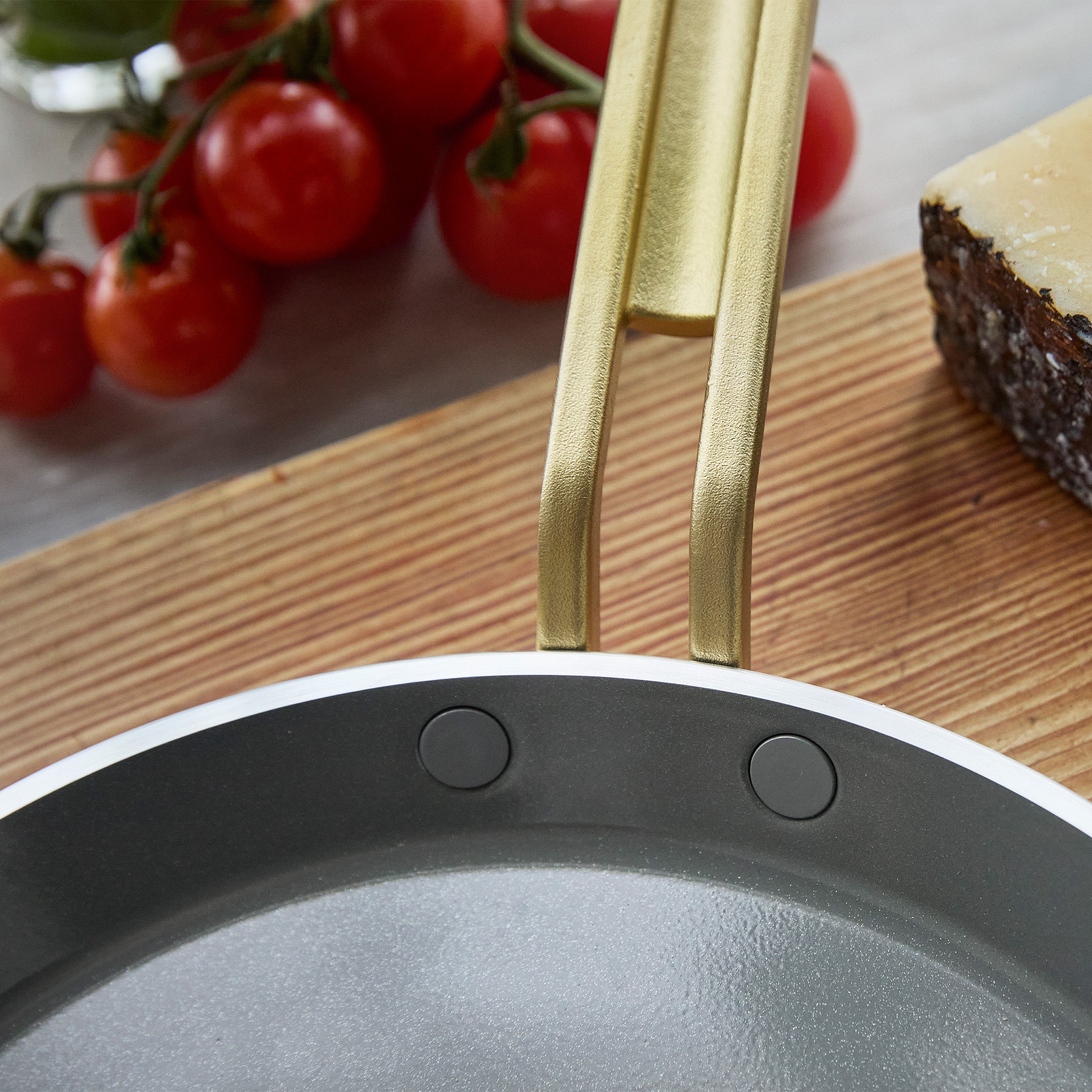 Gold crepe pan handle on a wooden surface with tomatoes and cheese in the background