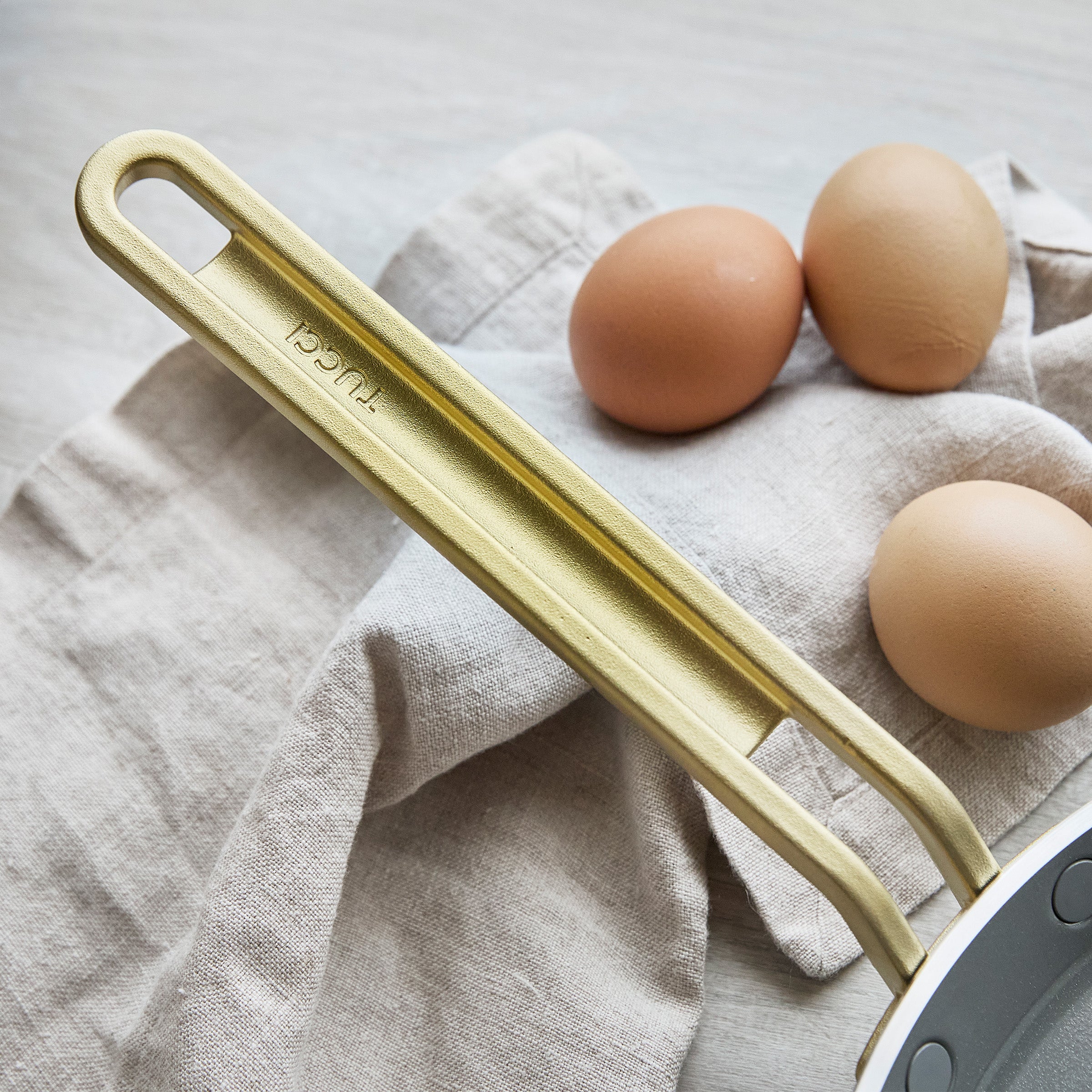 Gold crepe pan handle on a light gray towel with three brown eggs.
