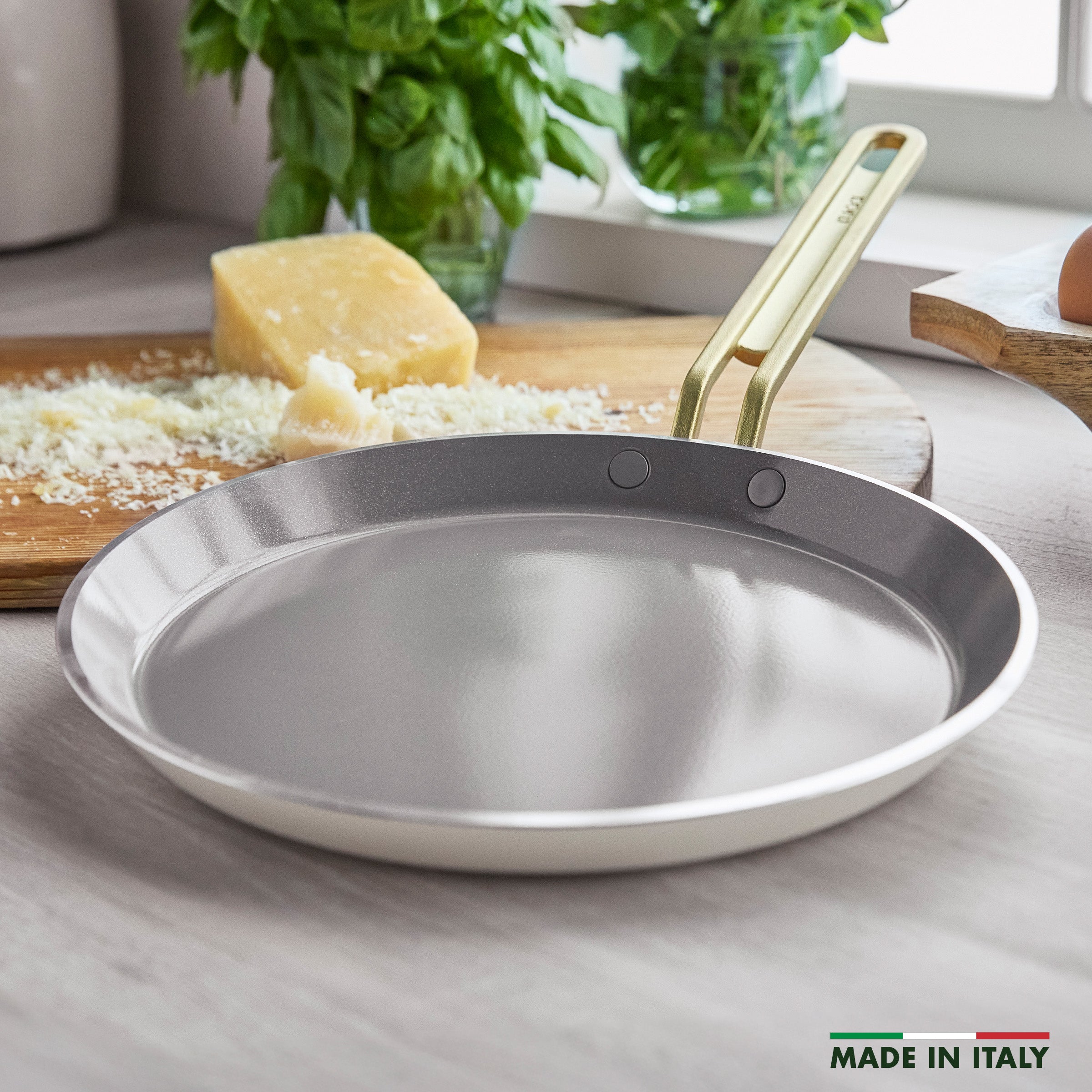 White crepe pan with a gold handle on a kitchen counter with cheese and herbs.