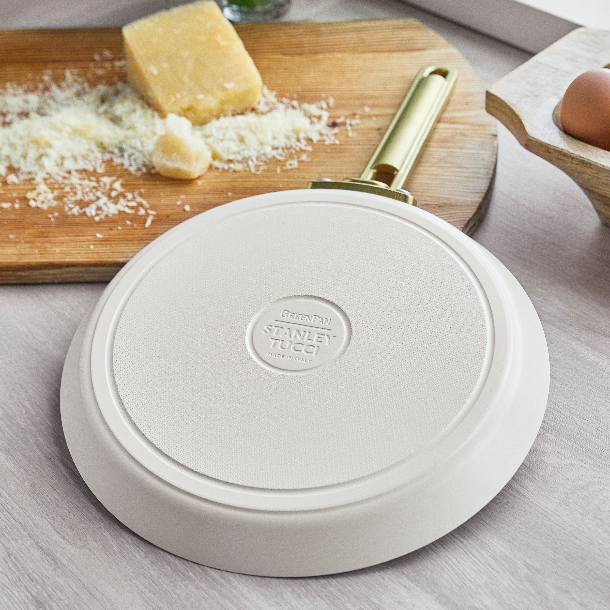 White crepe pan with 'Stanley Tucci' branding on a wooden surface with cheese and eggs.