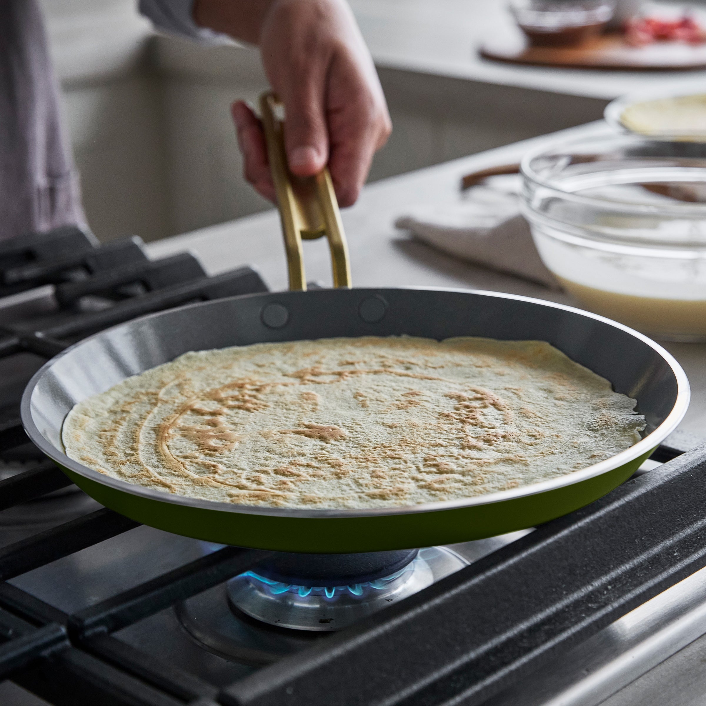 Person cooking a crepe in a green pan over a gas stove.