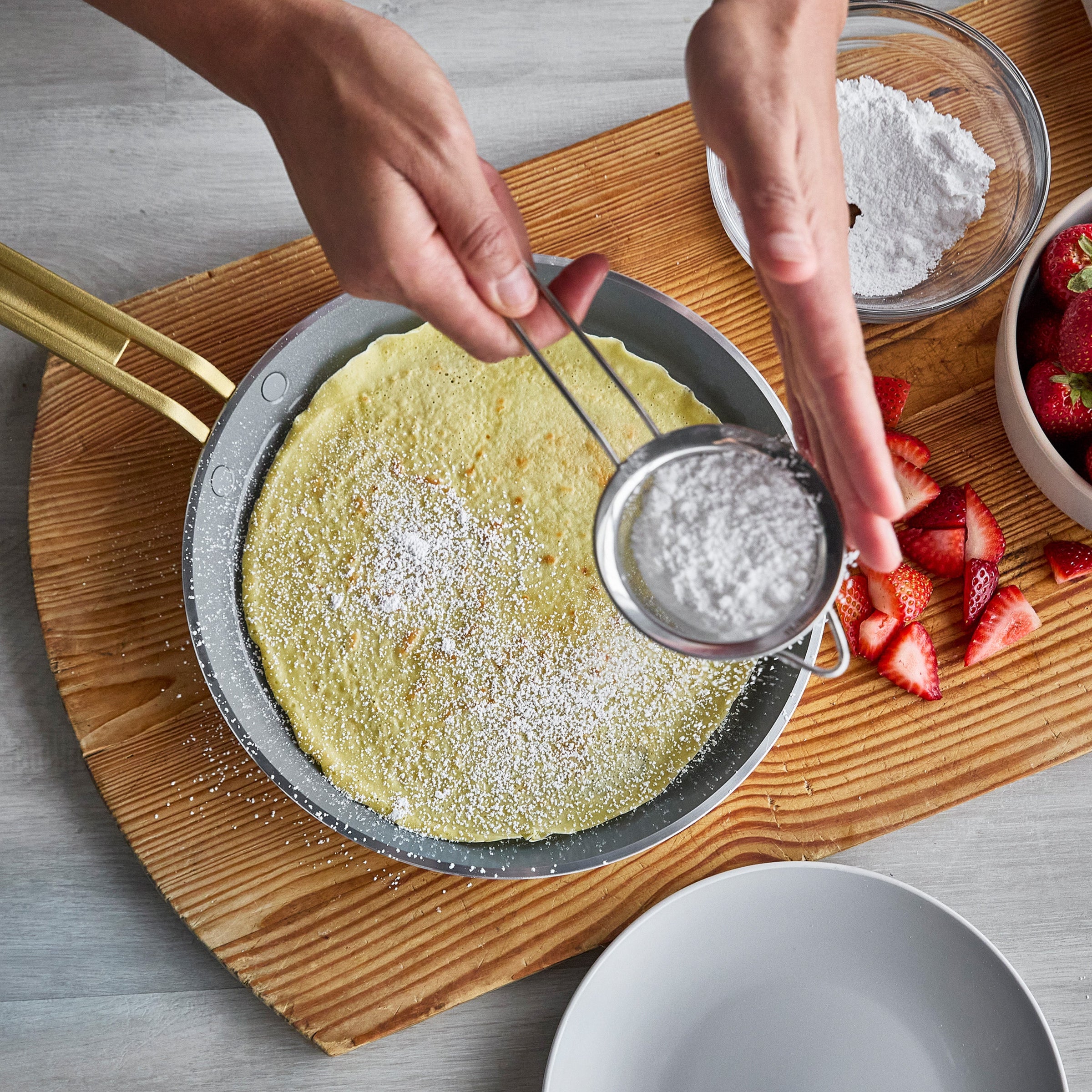 Person dusting a crepe with powdered sugar using a sifter on a wooden board.