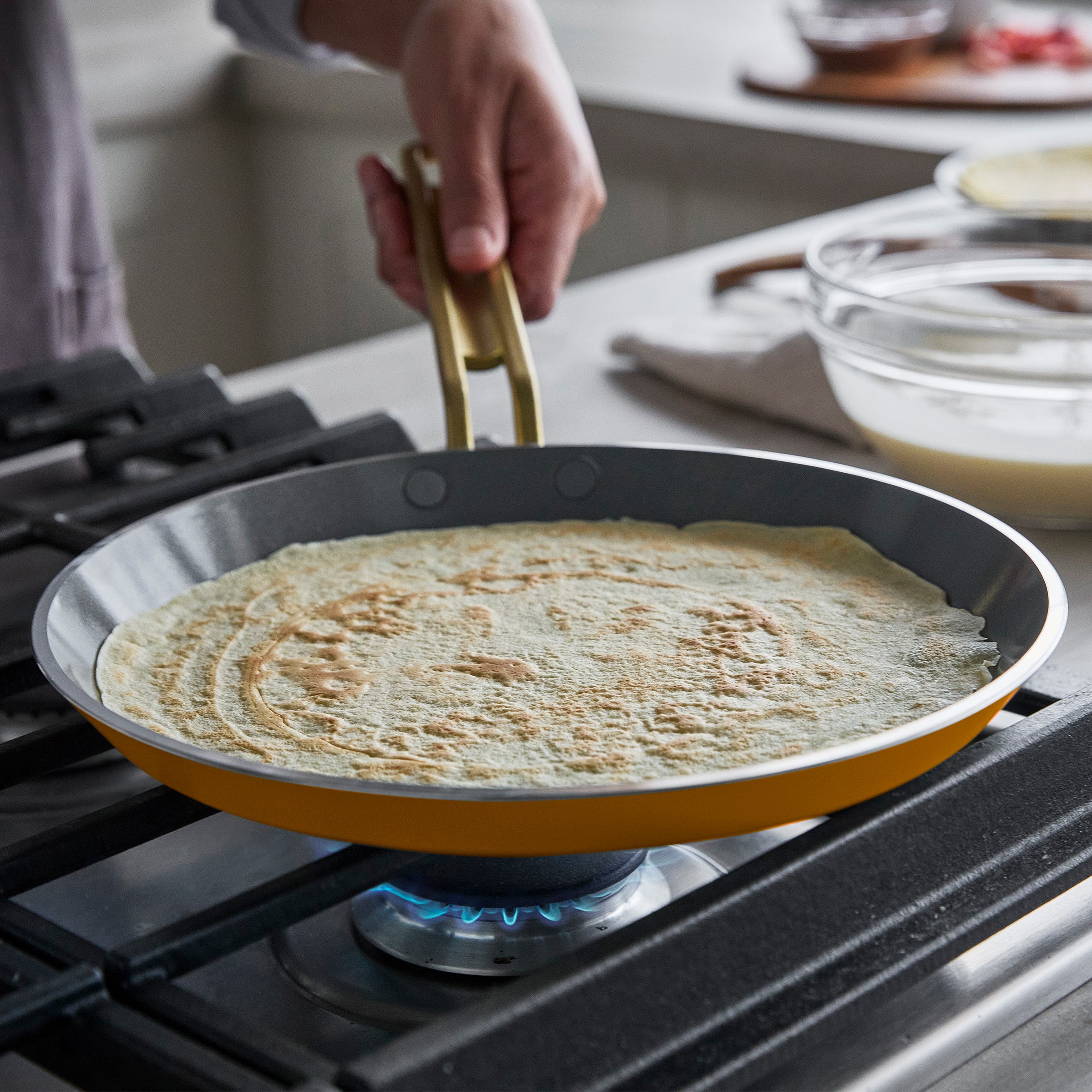 Person cooking a crepe in a yellow crepe pan on a gas stove.