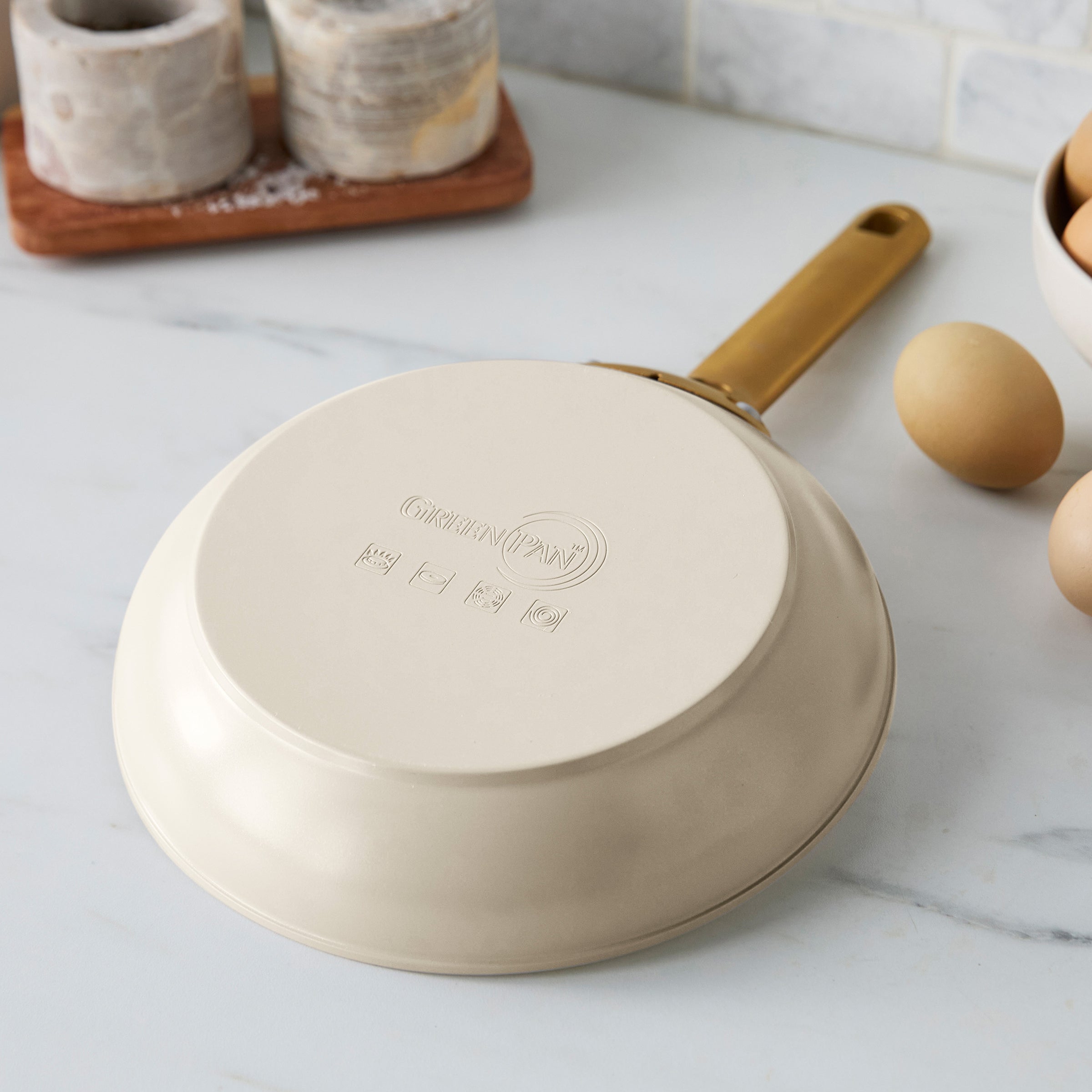 Taupe frying pan on a marble countertop with eggs and a wooden tray in the background
