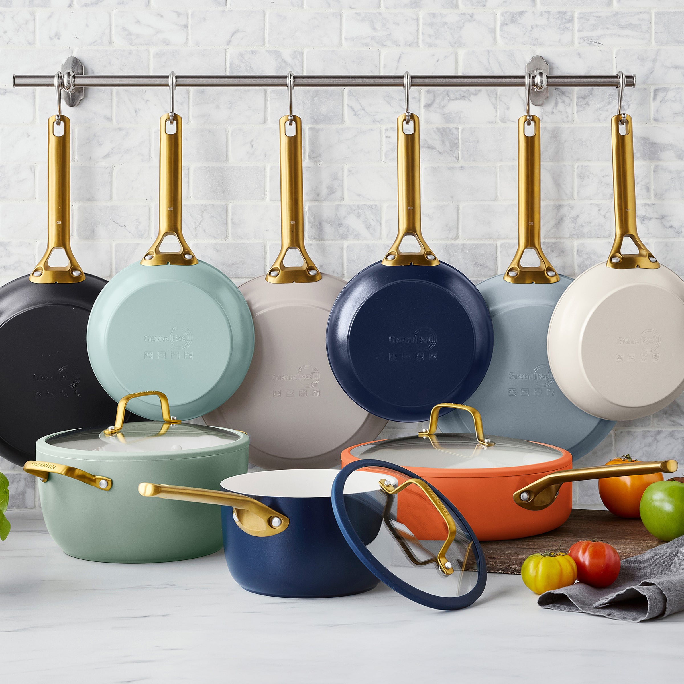Colorful cookware set with gold handles hanging on a rack against a white tiled wall.