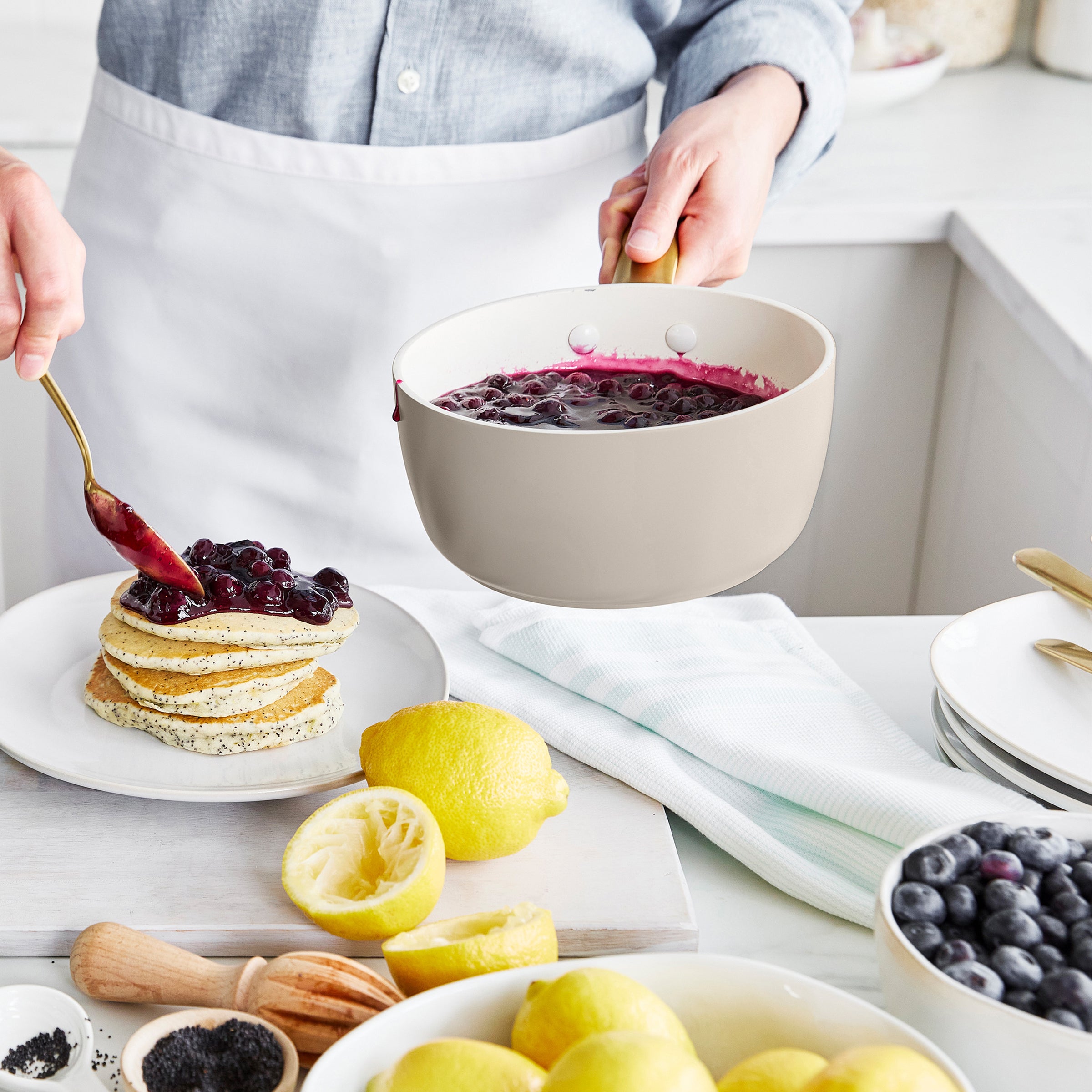 Person in a kitchen preparing pancakes with blueberry sauce, surrounded by lemons and blueberries.