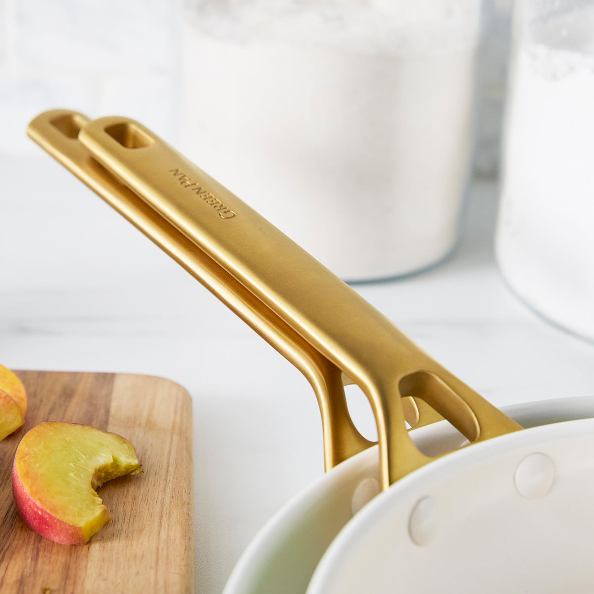 Gold frypan handles on a white surface with a wooden cutting board and apples.