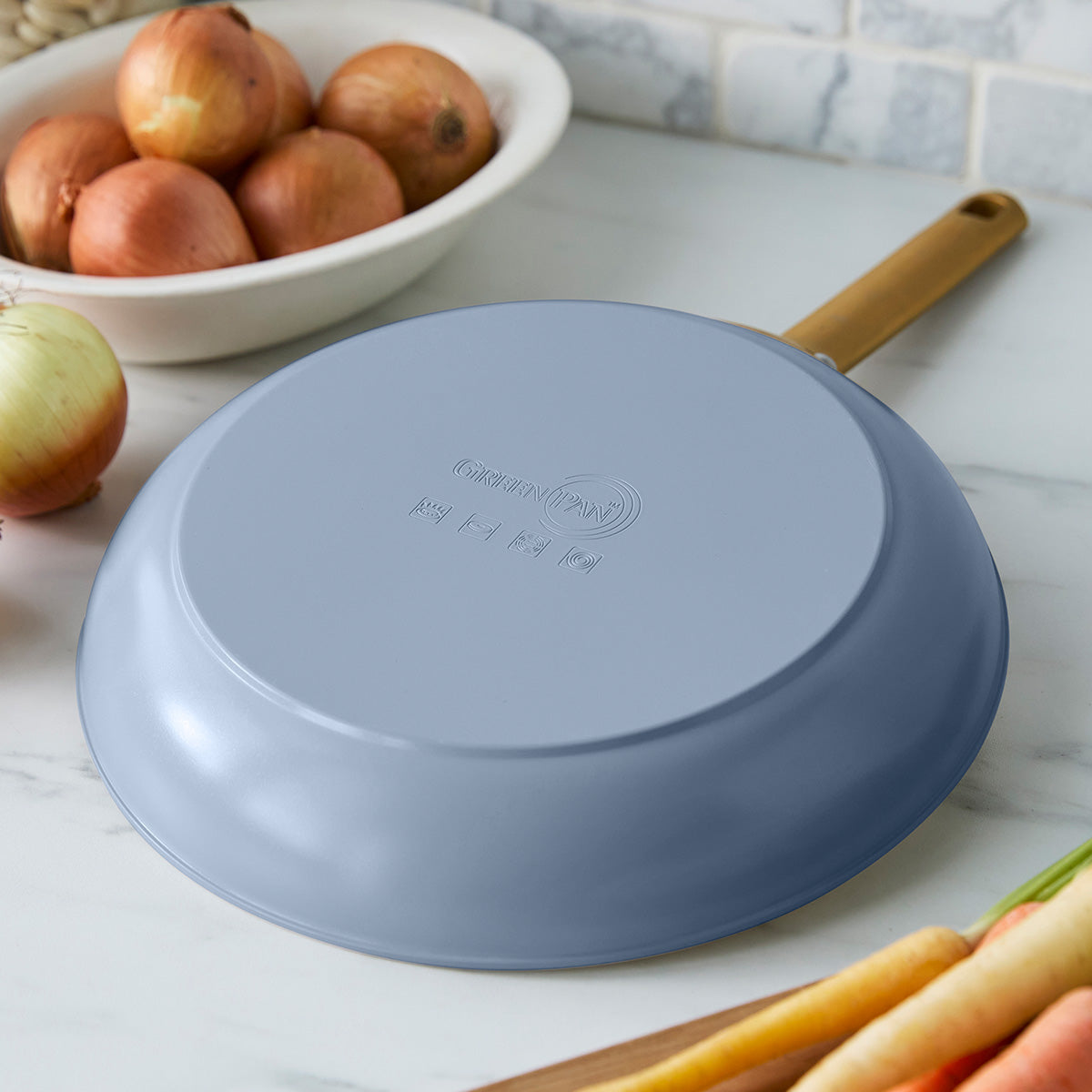 Blue frying pan on a kitchen counter with onions and carrots in the background
