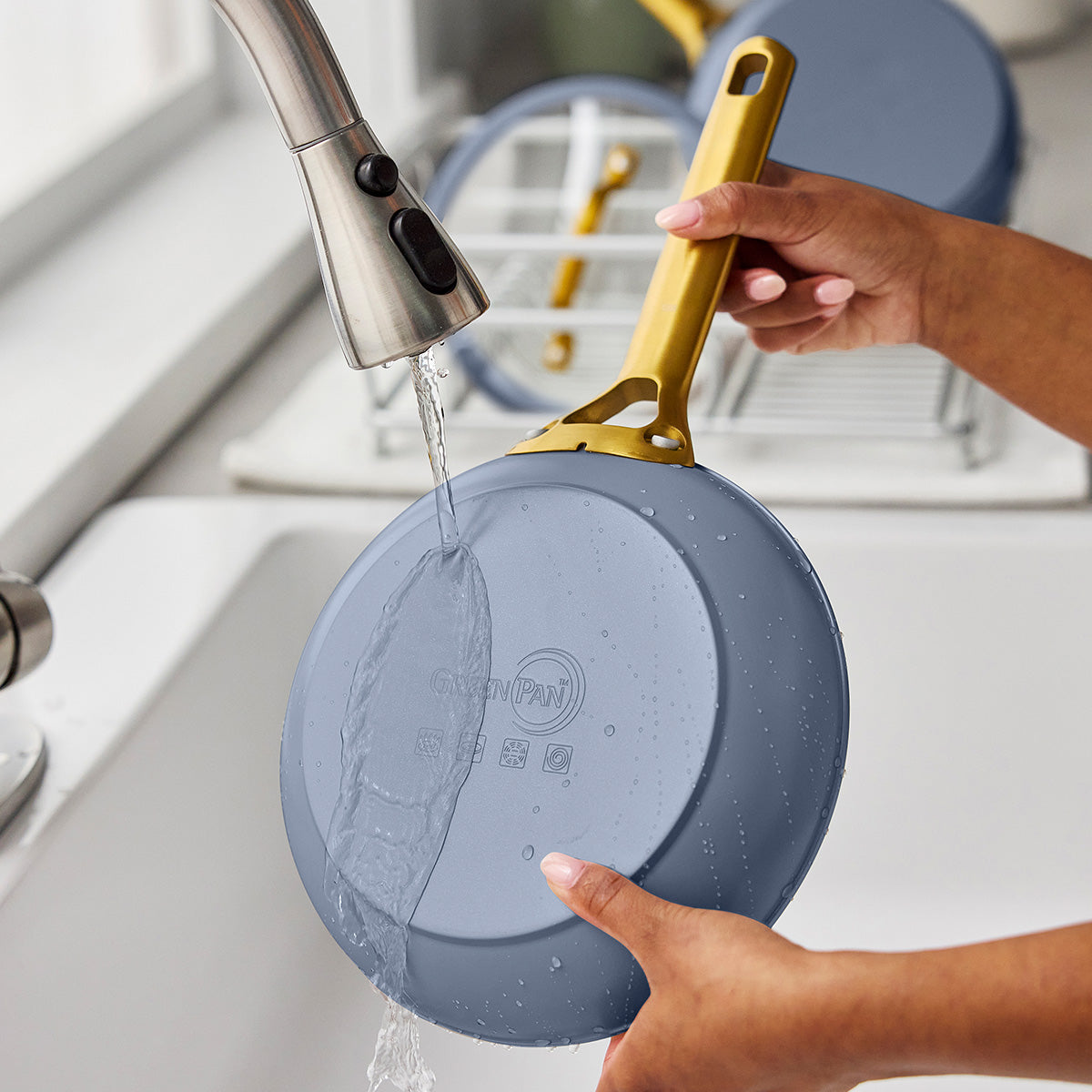 Person washing a blue frying pan with a gold handle under running water in a kitchen sink.