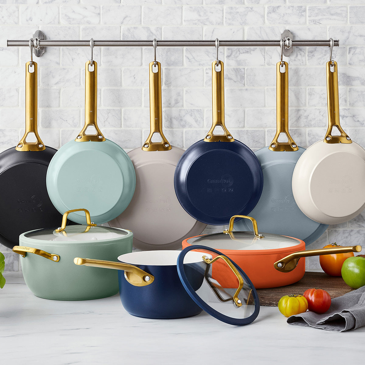 Colorful cookware set with gold handles hanging on a rack against a white tiled wall.