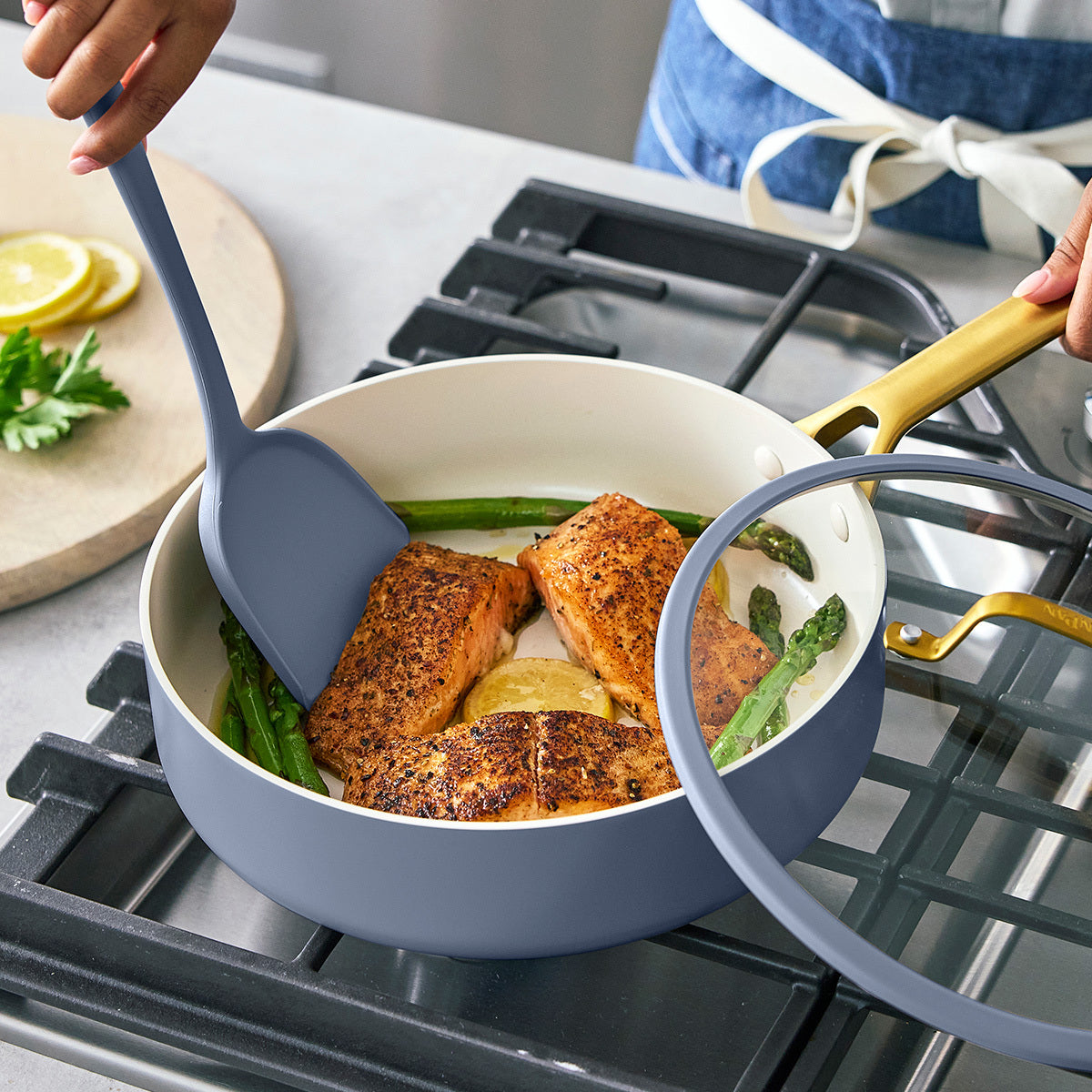 Person cooking salmon and asparagus in a blue saute pan on a stove with a spatula.