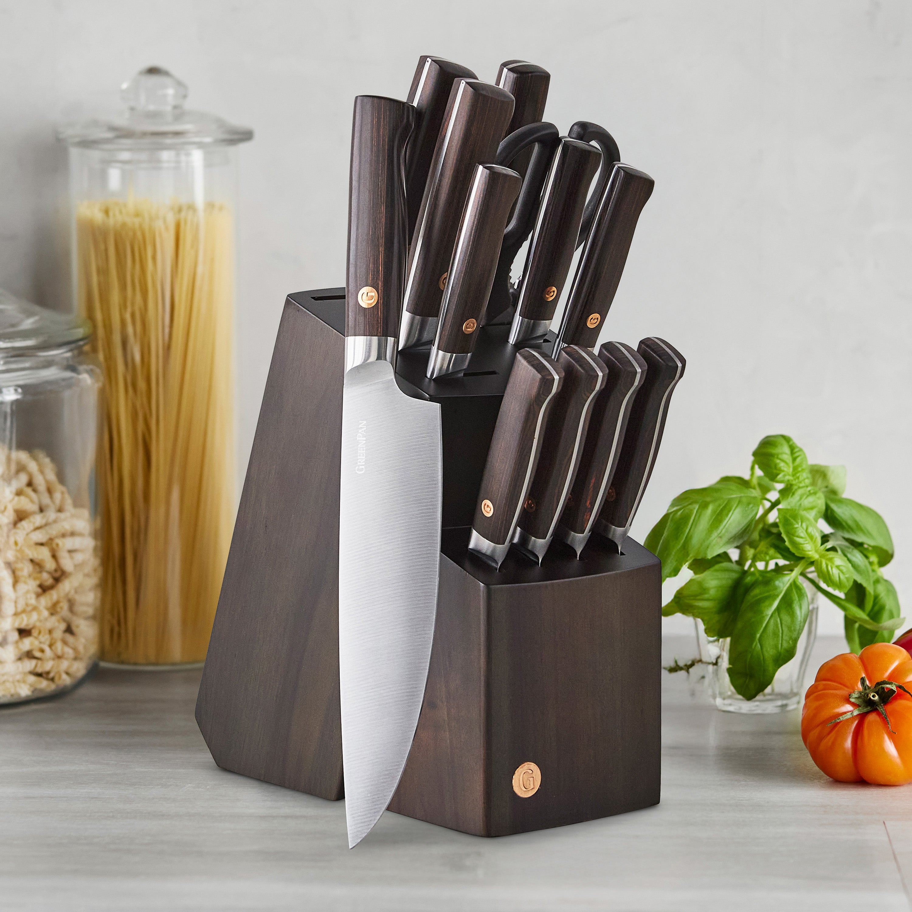 Set of knives in a knife block on a kitchen counter with pasta and basil in the background.