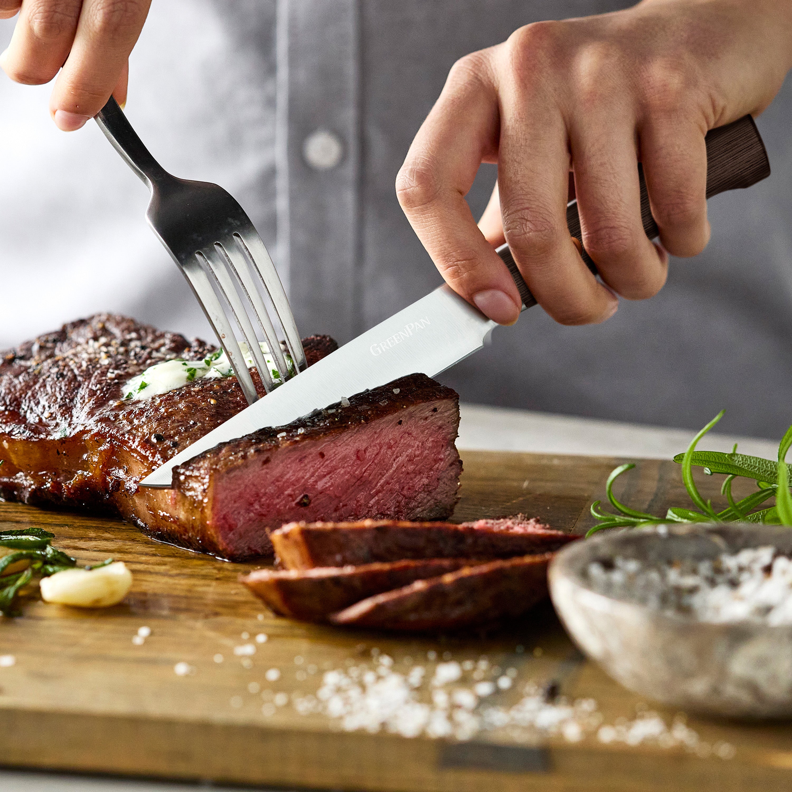 Person cutting a steak with a knife and fork on a wooden cutting board.