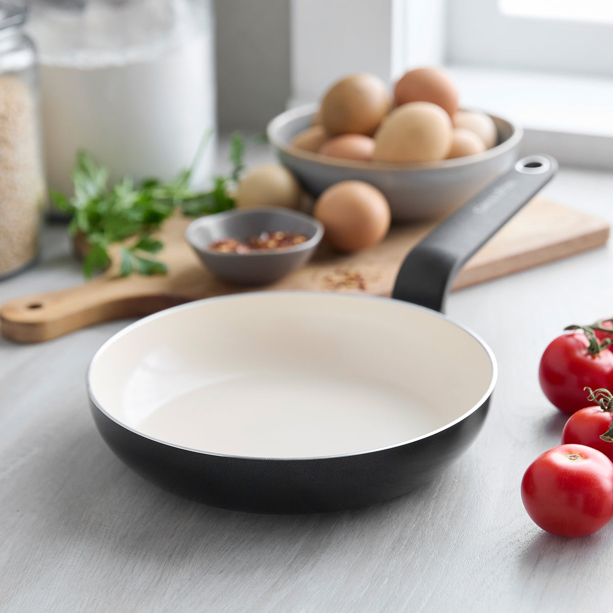 Black ceramic 8-inch frypan on a countertop, with eggs, tomatoes, and herbs visible in the background.