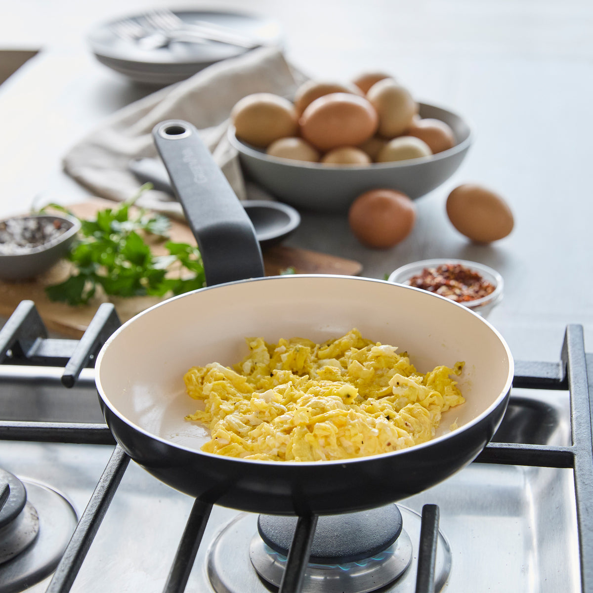 Black ceramic 8-inch frypan with scrambled eggs, fresh eggs, and herbs visible in the background on a stovetop.