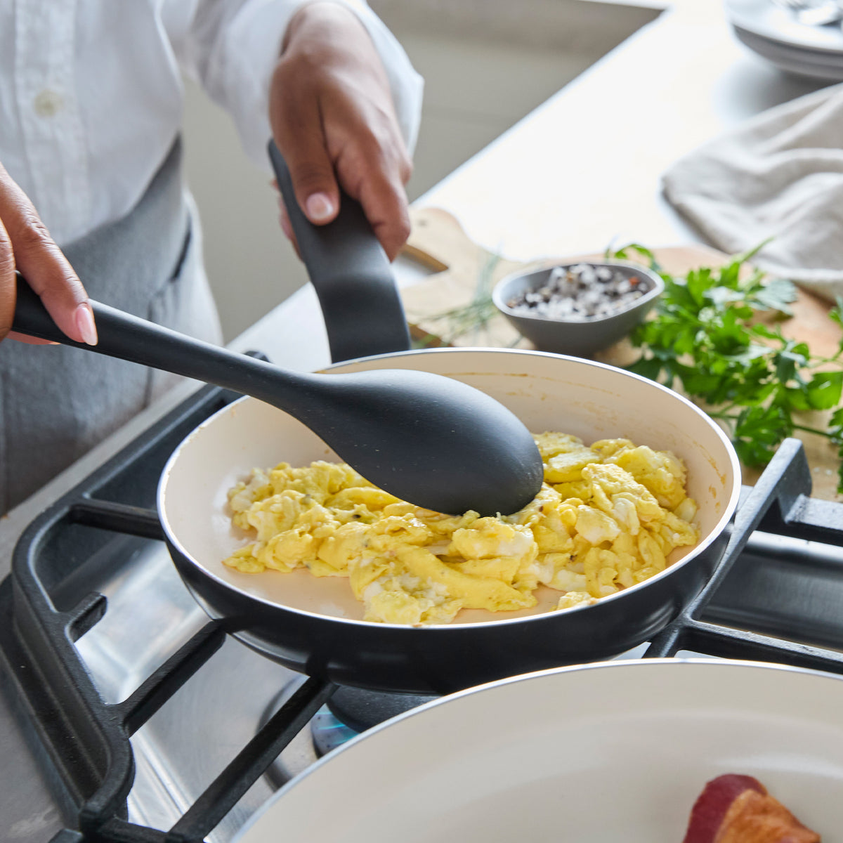 Scrambled eggs in a black ceramic nonstick frypan with black utensils; fresh herbs on the side.