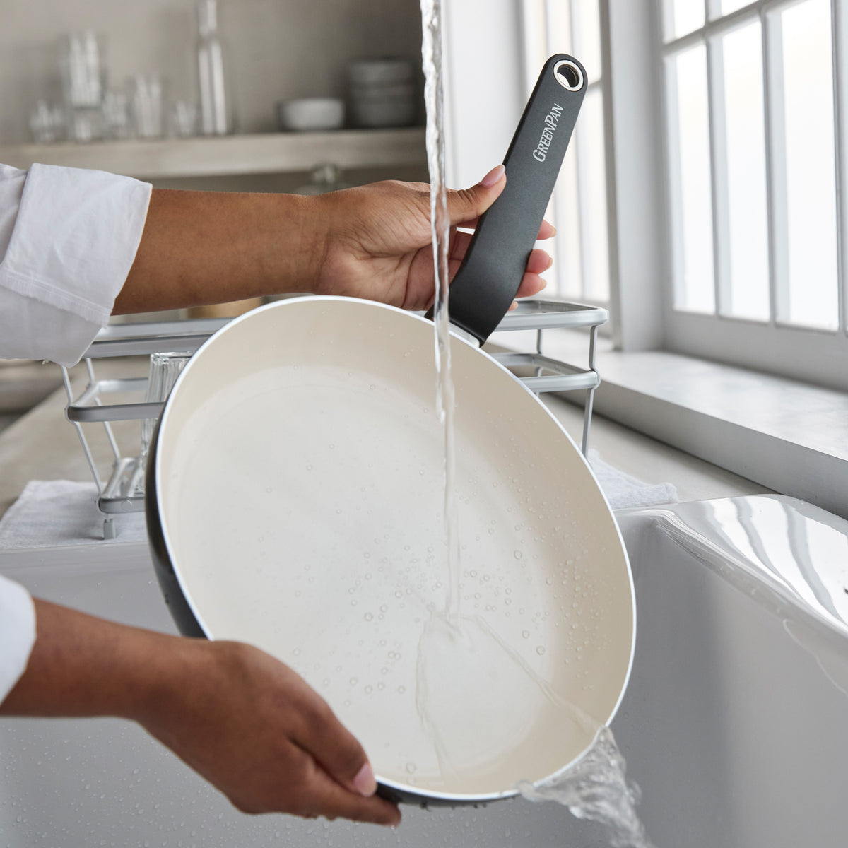 Black ceramic nonstick 8-inch frypan being rinsed under running water in a kitchen sink by a window.