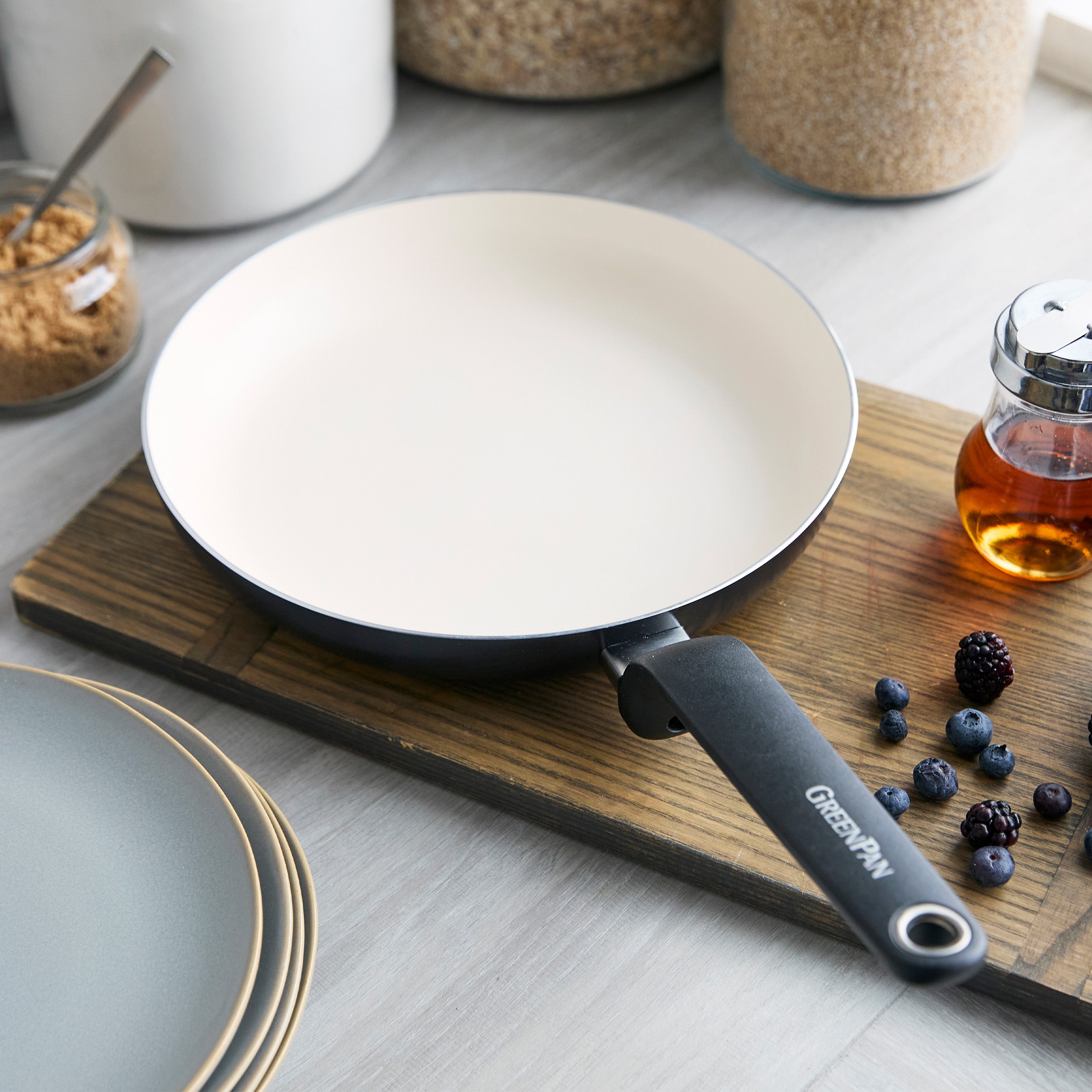 Black ceramic 8-inch frypan on a wooden surface near plates, berries, syrup bottle, and jars of grains.
