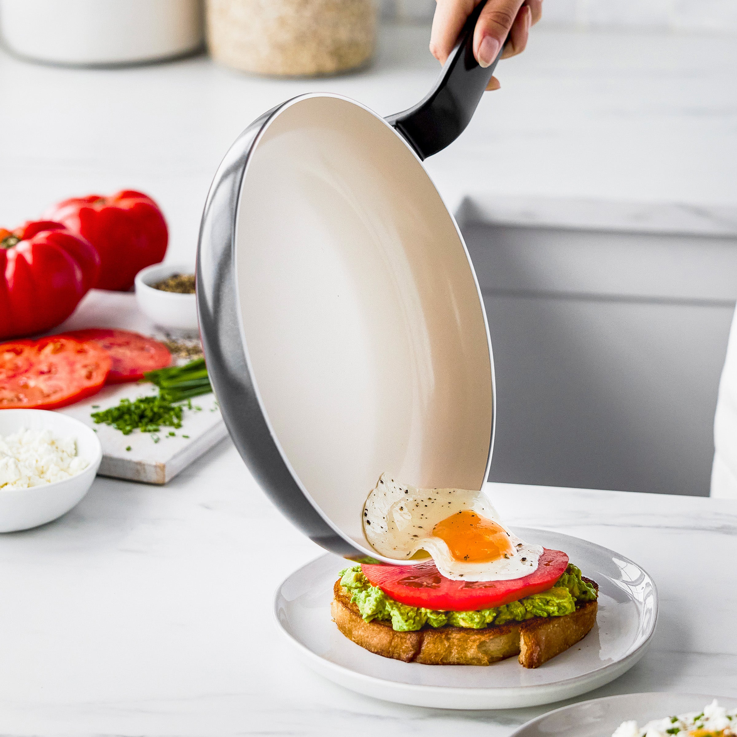 A hand slides a fried egg from a black ceramic 10-inch frypan onto avocado toast in a bright kitchen.