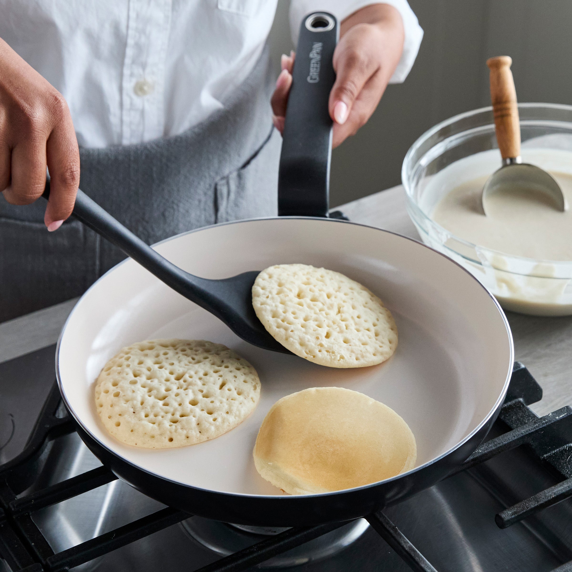 Black ceramic nonstick 8 frypan flipping pancakes, with glass bowl of batter and a ladle beside it.