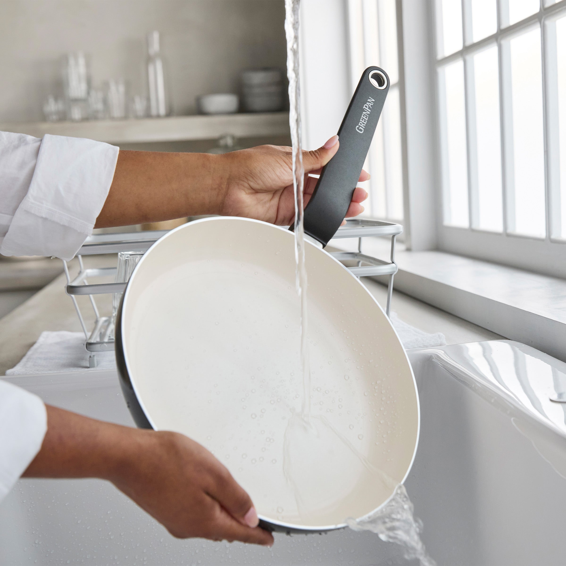 Black ceramic 10 frypan being rinsed under running water in a bright kitchen sink.