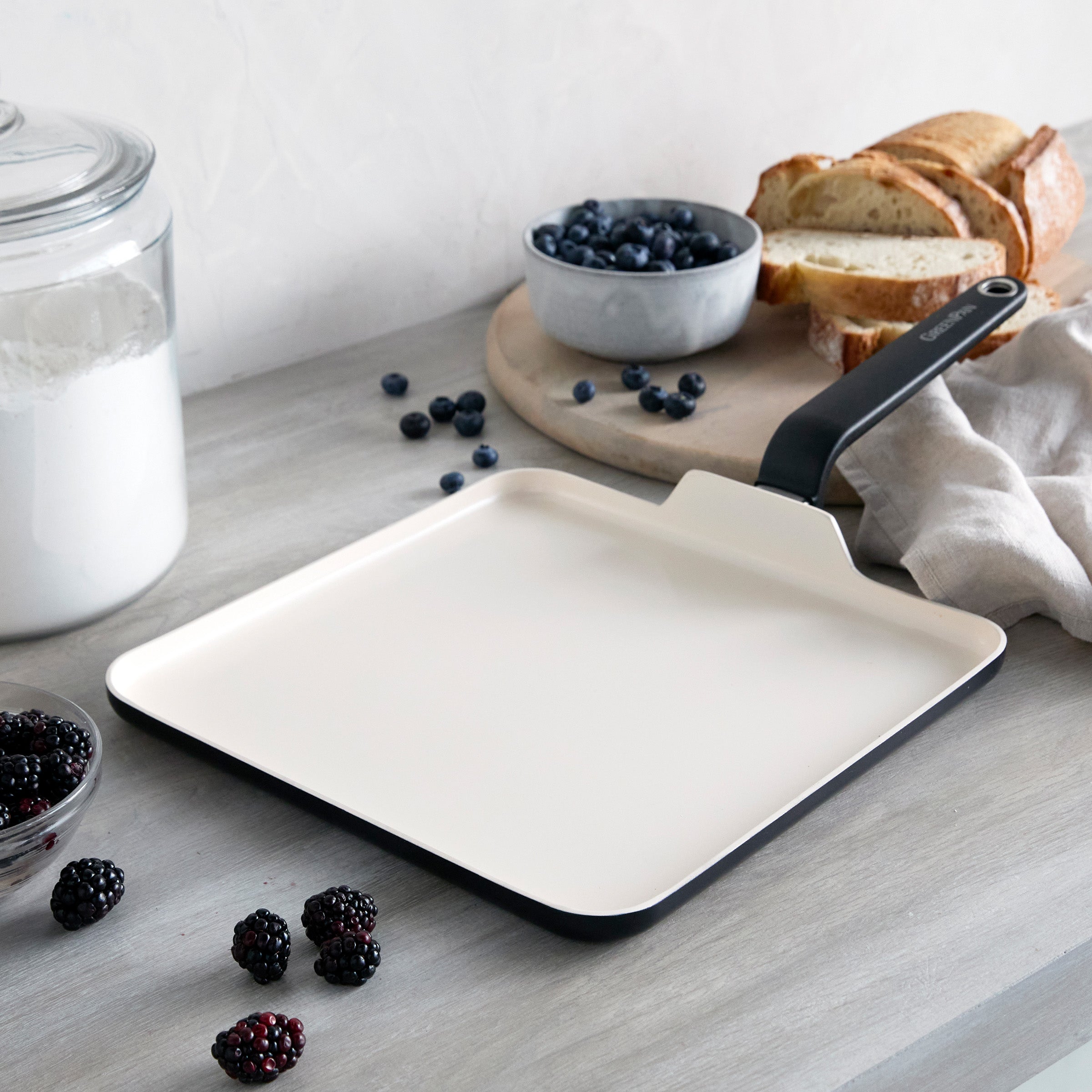 Black square ceramic griddle on a counter with bread, berries, flour, and a linen cloth.