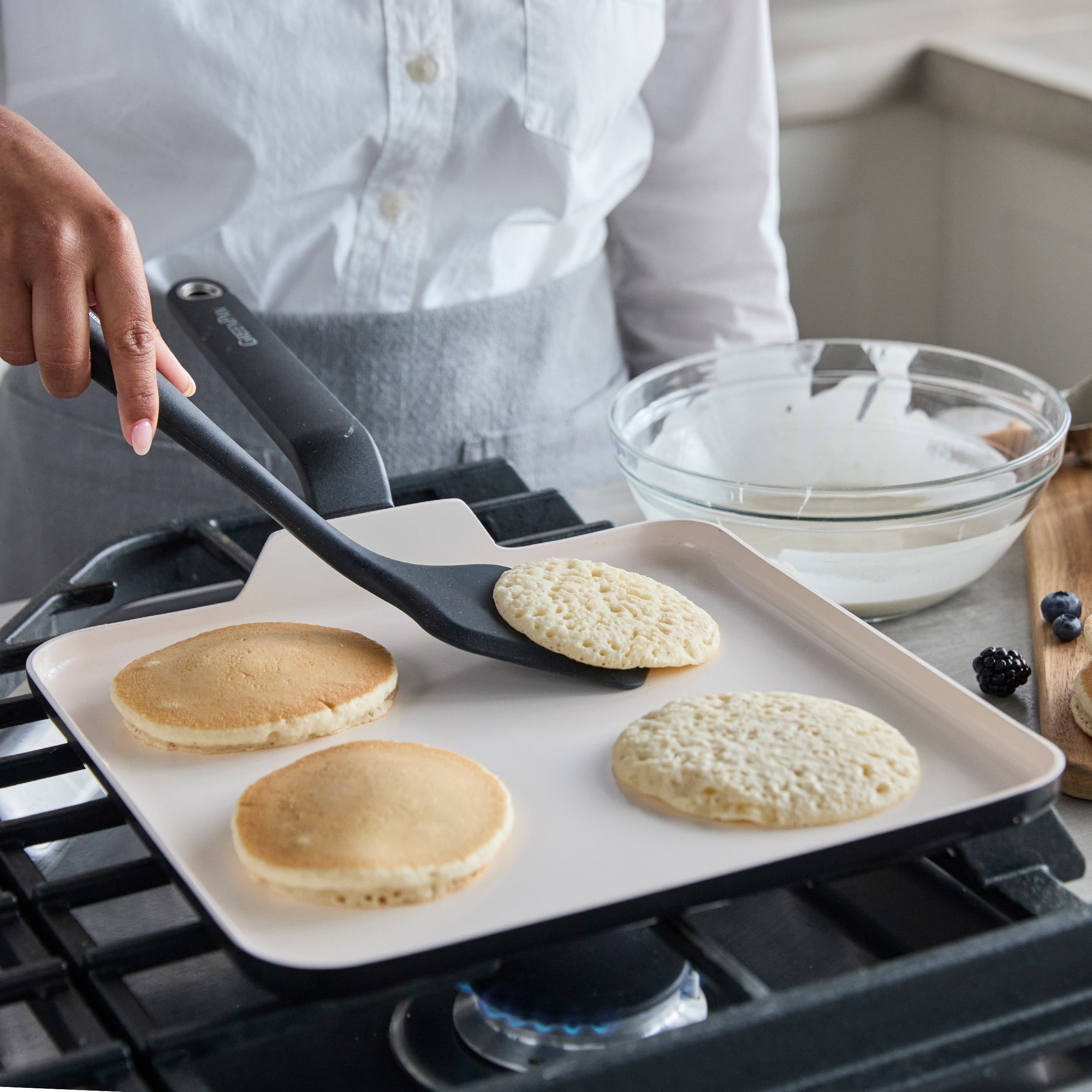 Square ceramic griddle cooking pancakes, with bowls of batter and berries visible in the background.