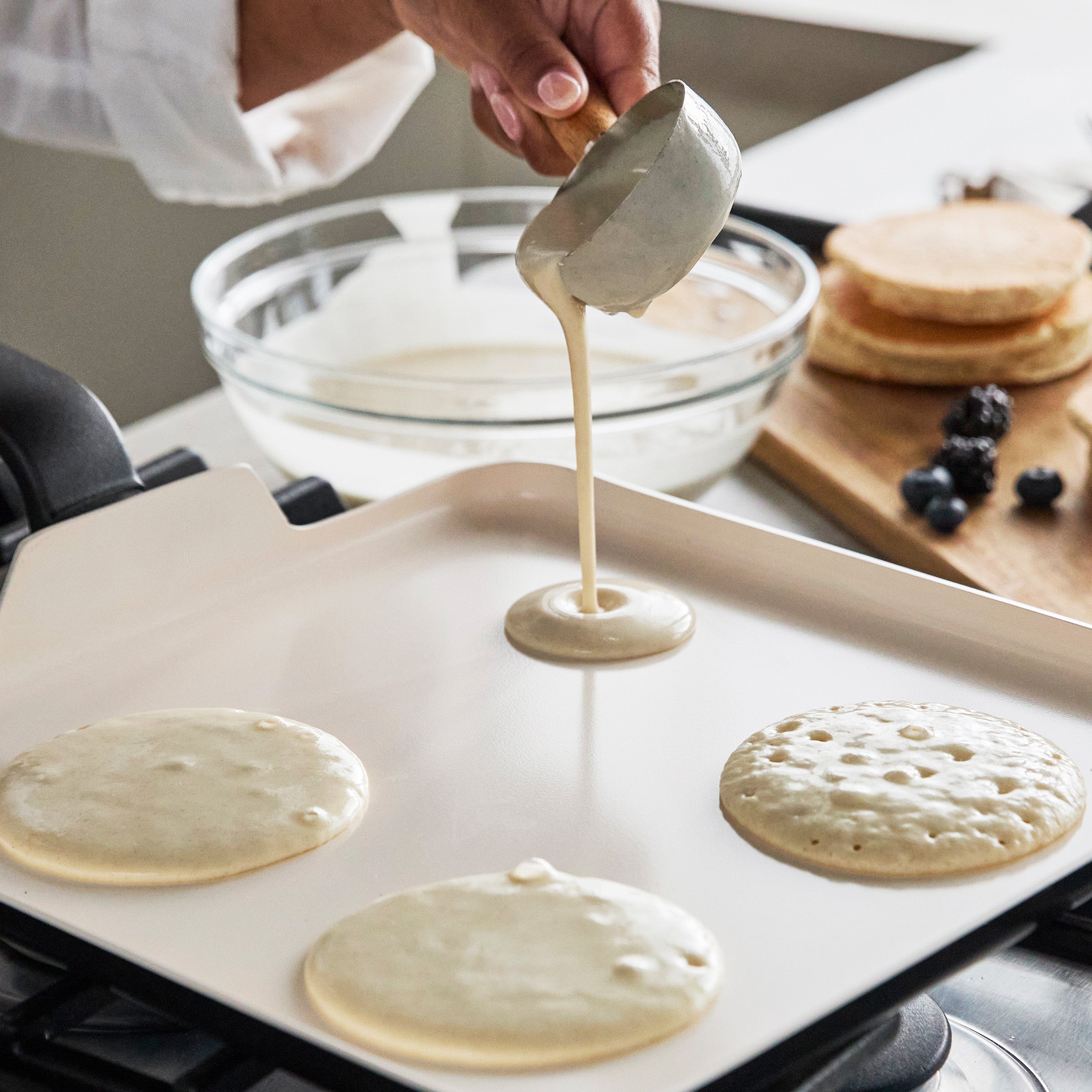 Batter being poured onto a square ceramic griddle with pancakes, a bowl of batter, and fruit visible nearby.