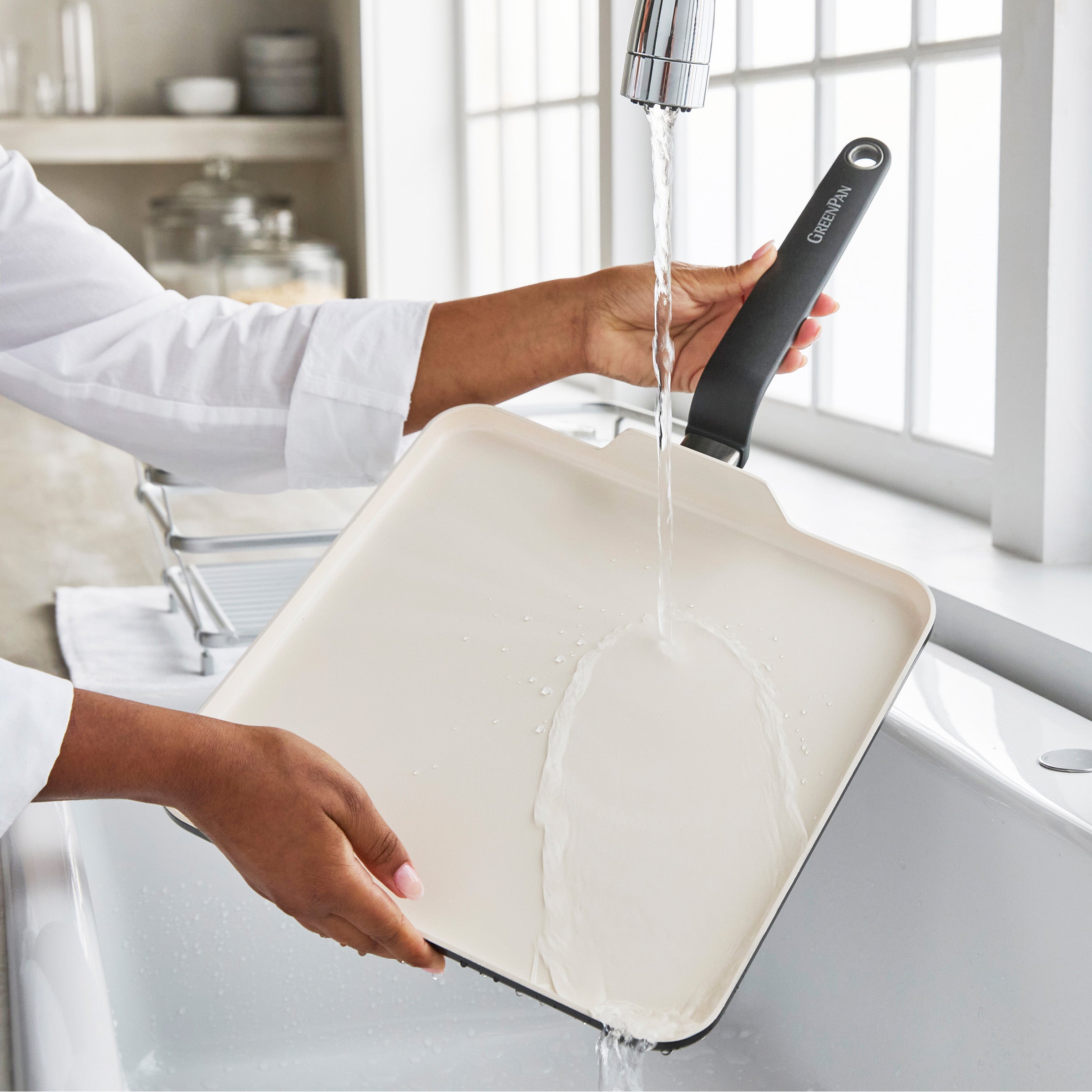 Black ceramic nonstick square griddle being rinsed under running water in a bright kitchen sink.